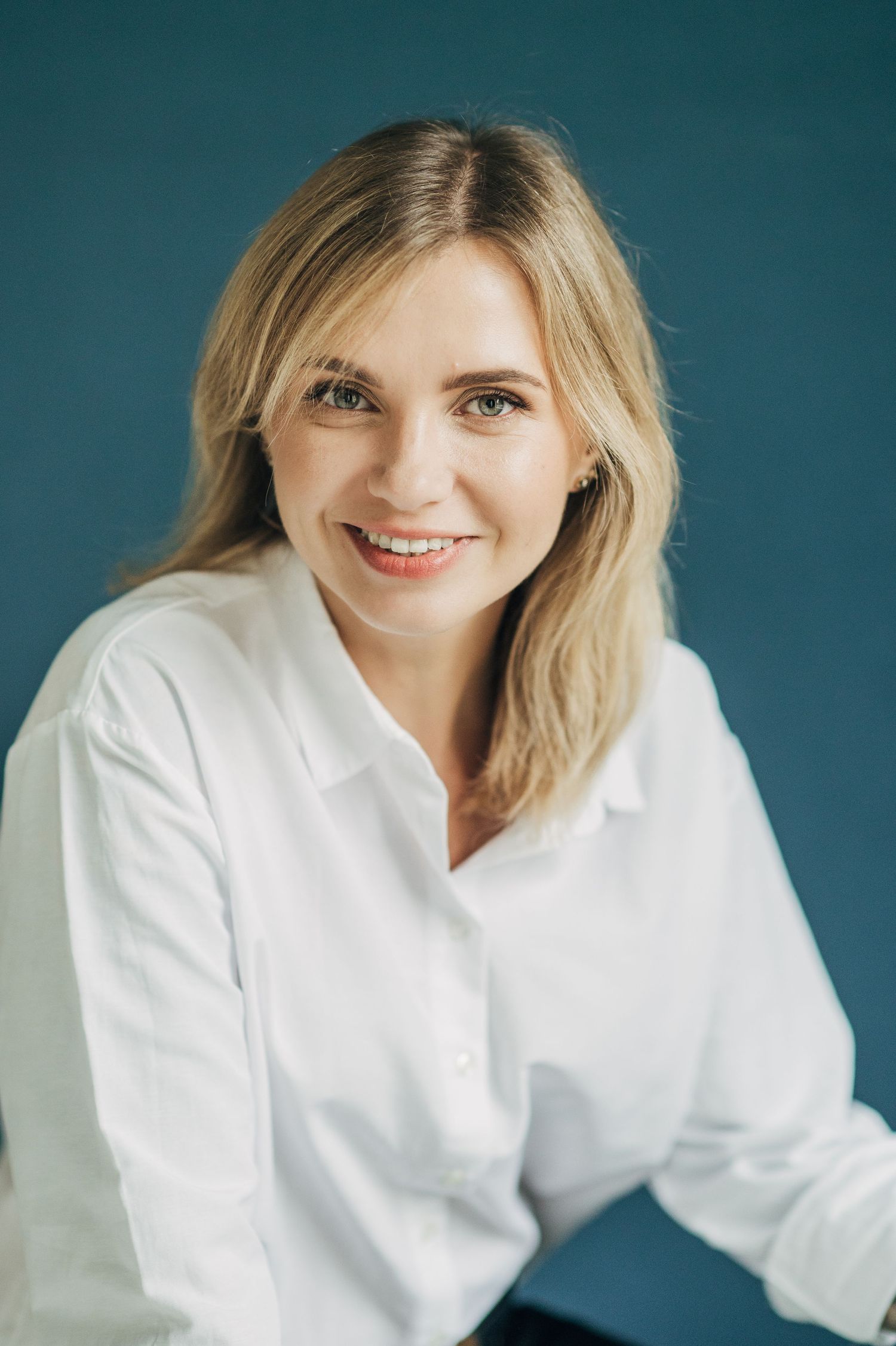 Professional headshot in white button-down shirt against teal background with natural smile.