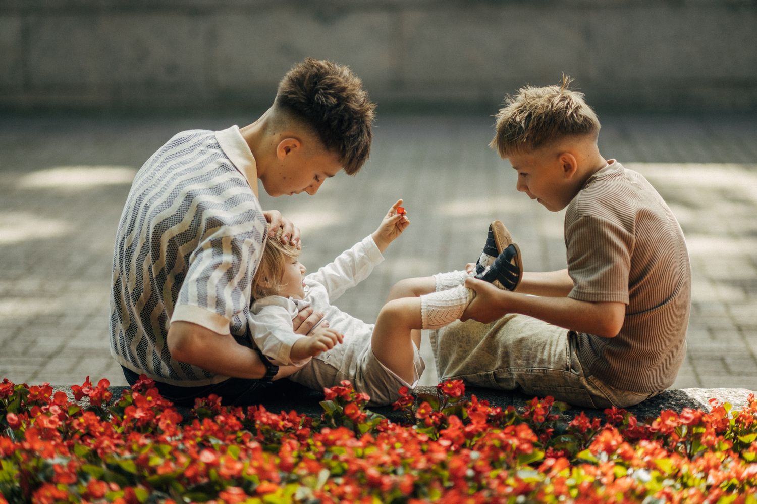 Two teenage boys sitting among red flowers, gently playing with a baby outdoors.