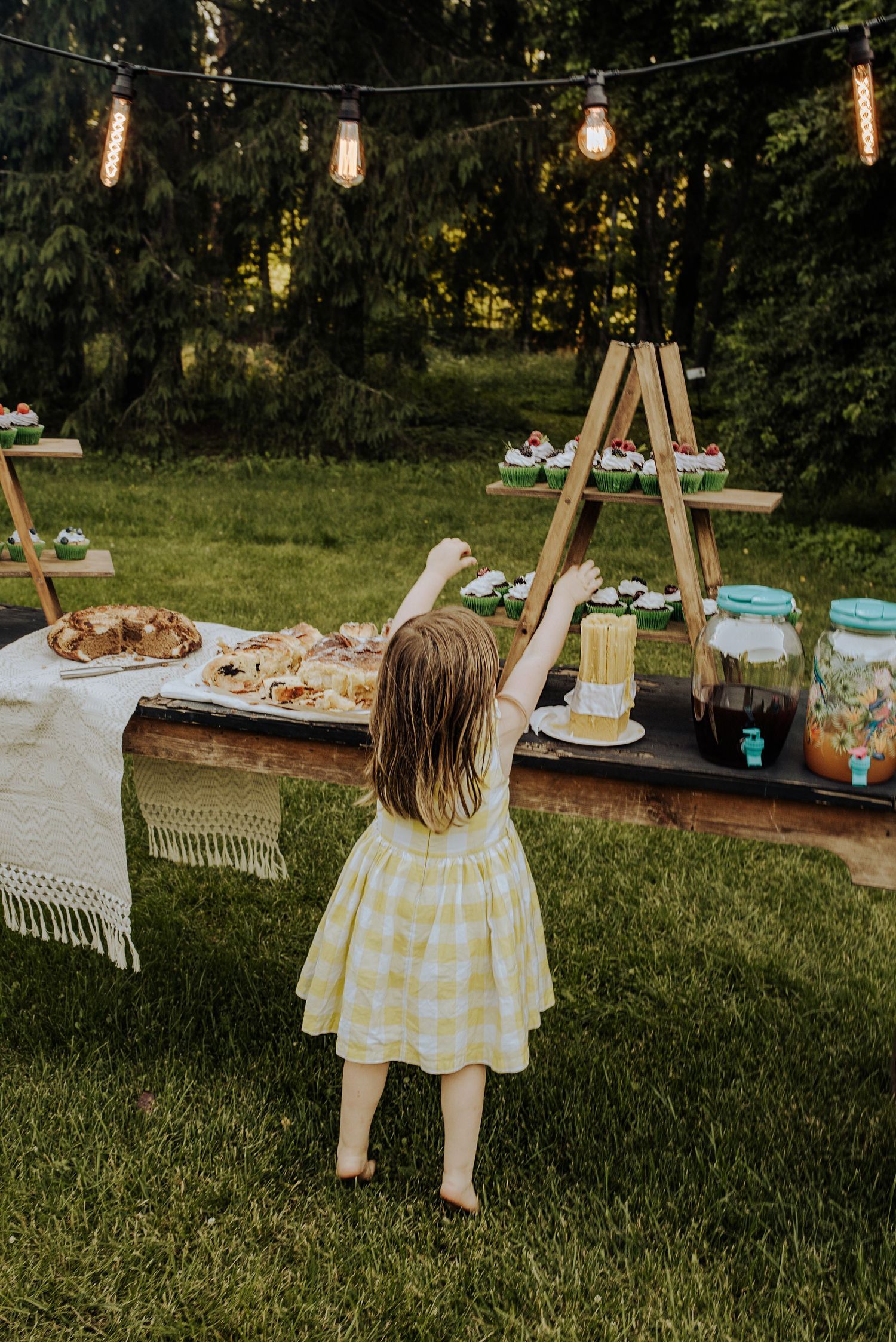 Little girl in yellow dress reaching for treats at an outdoor party dessert table with string lights.