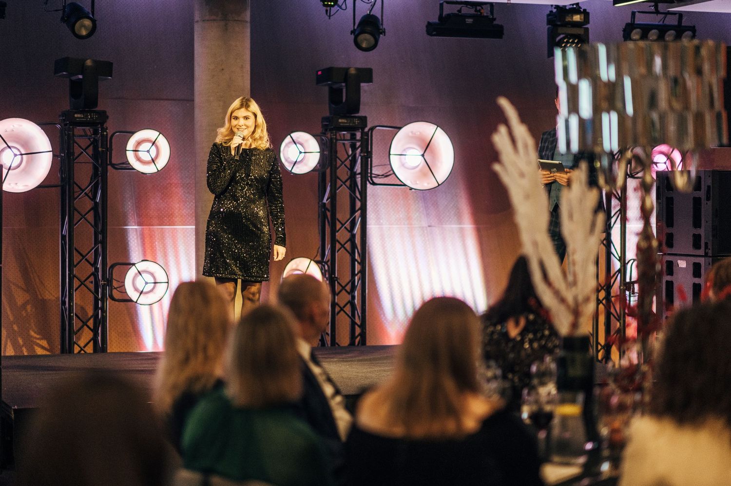 A performer in a black dress stands on stage with illuminated circular lights in the background as audience members watch.
