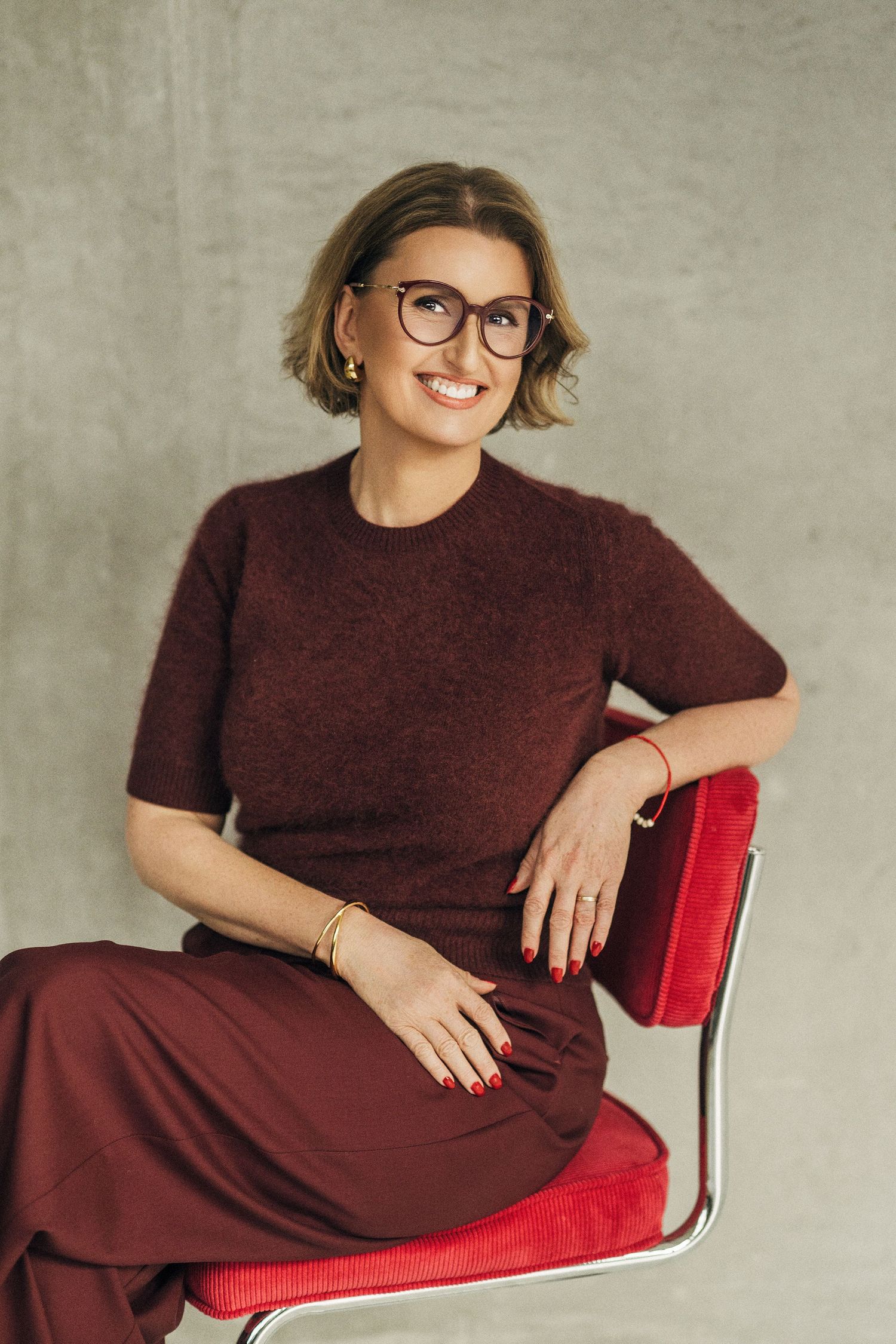 Person in burgundy dress and glasses sits comfortably on red upholstered chair against neutral wall.