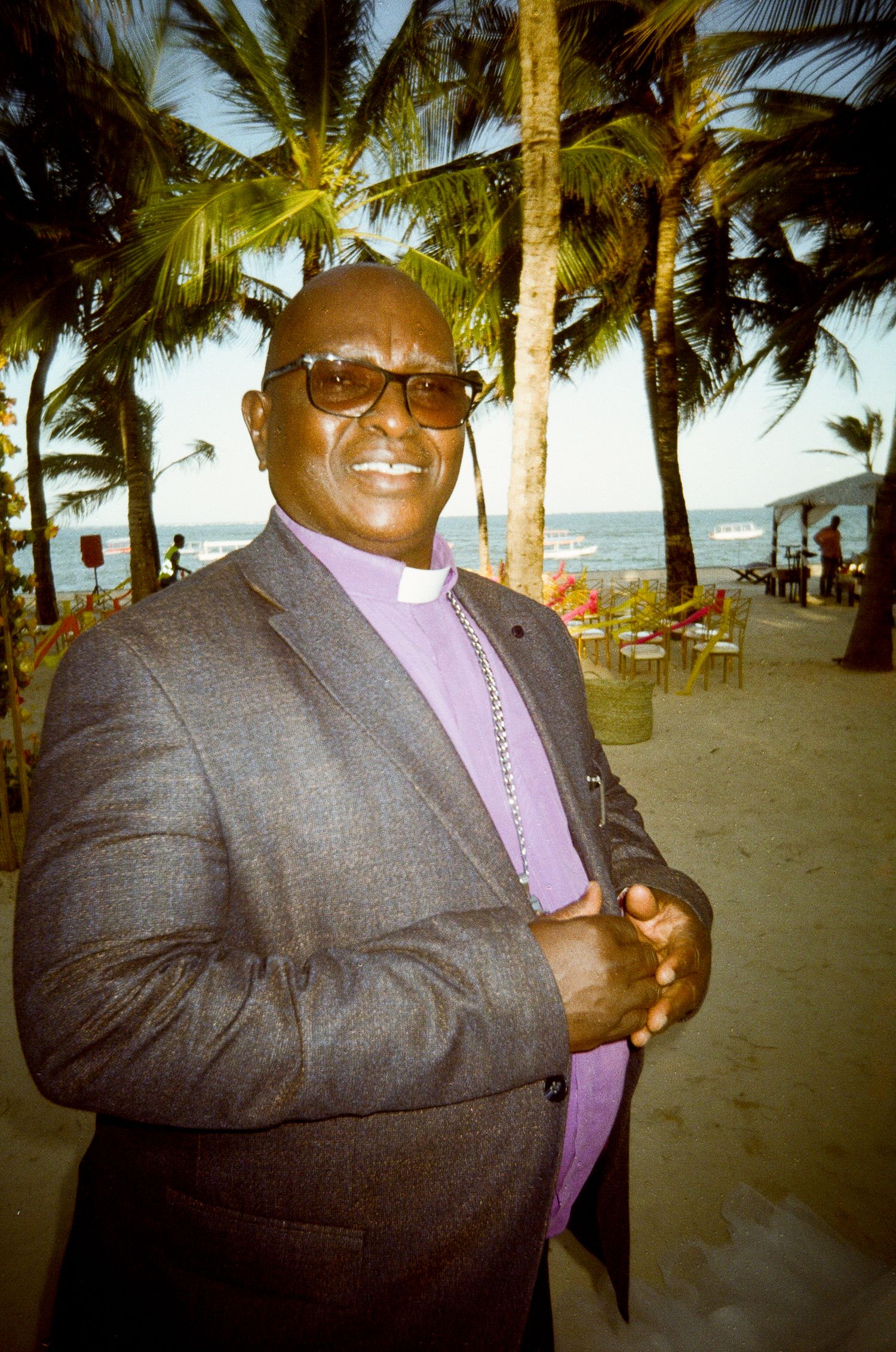 Person in a grey suit and purple shirt poses confidently on a palm-lined beach.