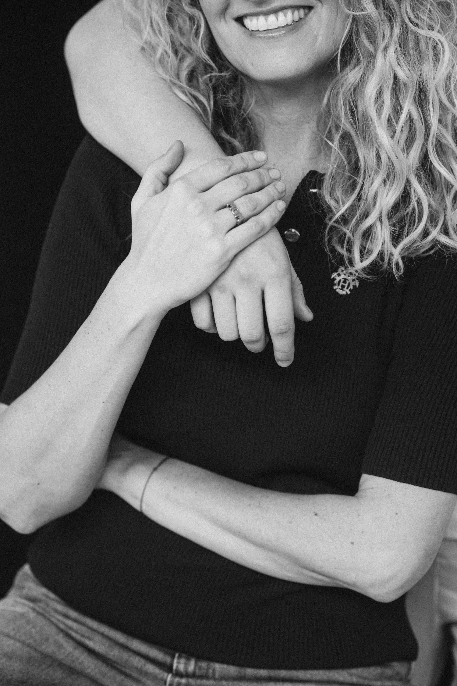 Black and white photo of a smiling woman with curly hair wearing a ring, being embraced from behind by a partner.