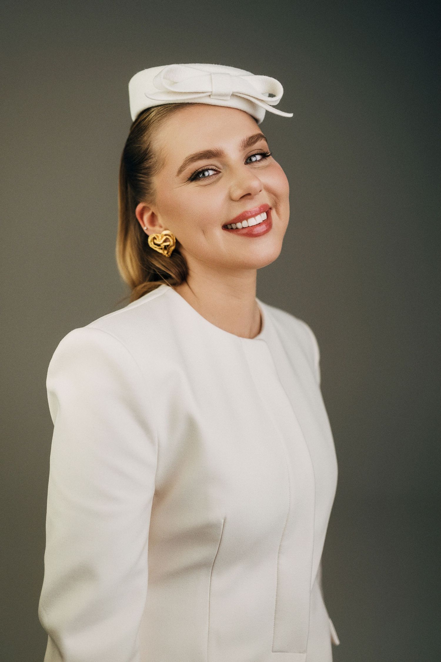 A person wearing a white baseball cap and white sweater smiles warmly against a neutral background.