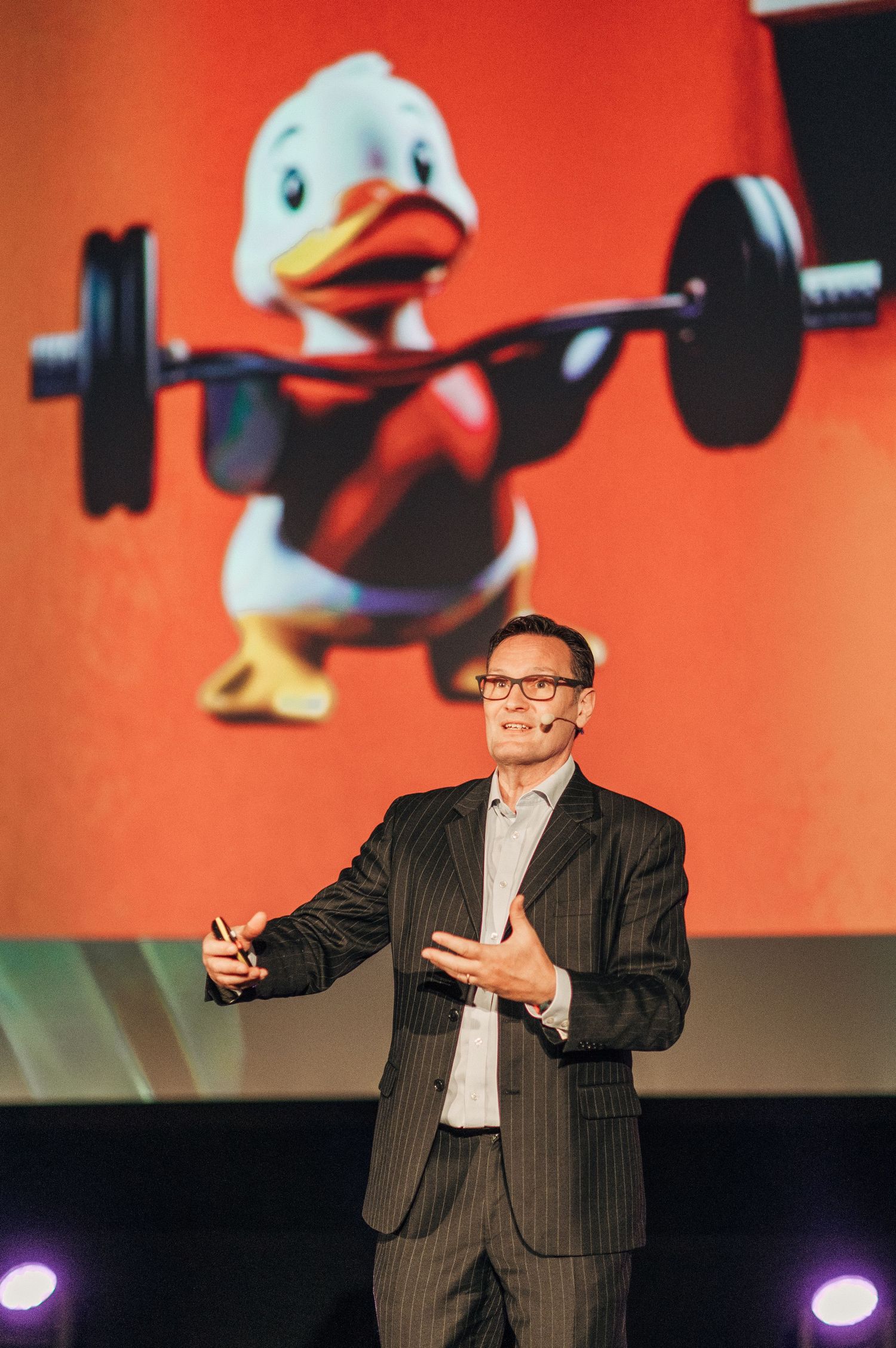 Speaker presenting on stage with animated graphics displayed on large red screen behind them.