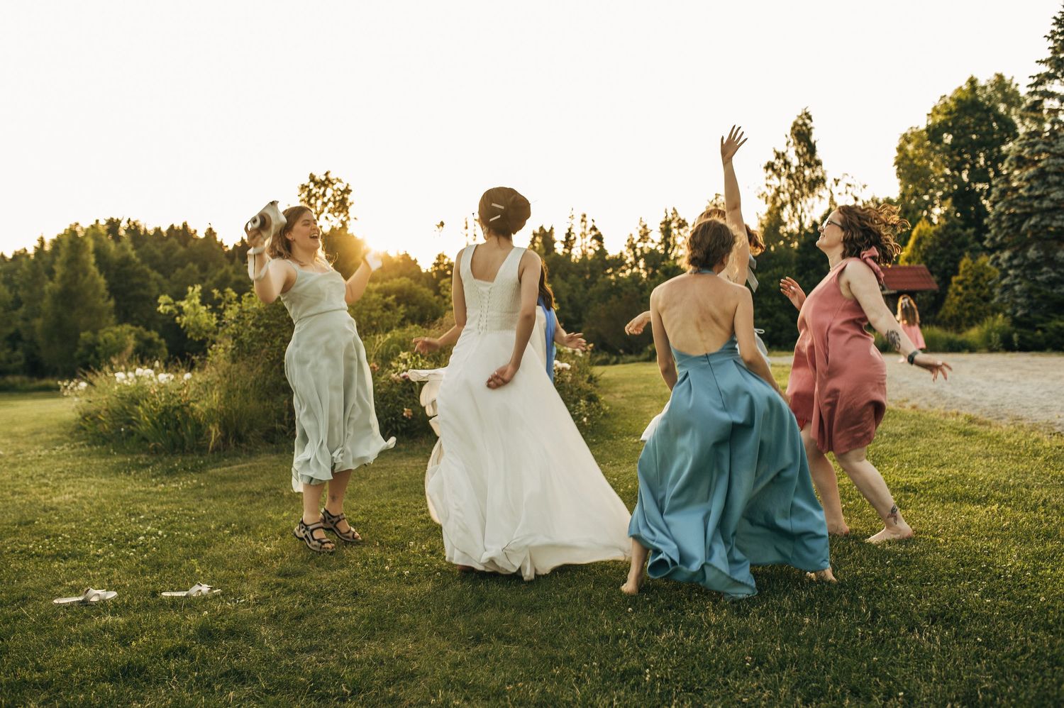 Bride and bridesmaids dancing joyfully outdoors at golden hour on lush green lawn at summer wedding.