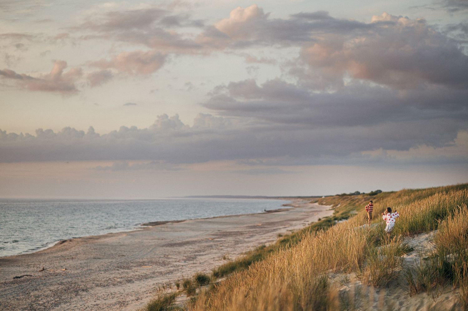 Couple walking along a serene sandy beach at sunset with dramatic clouds over calm ocean waters.