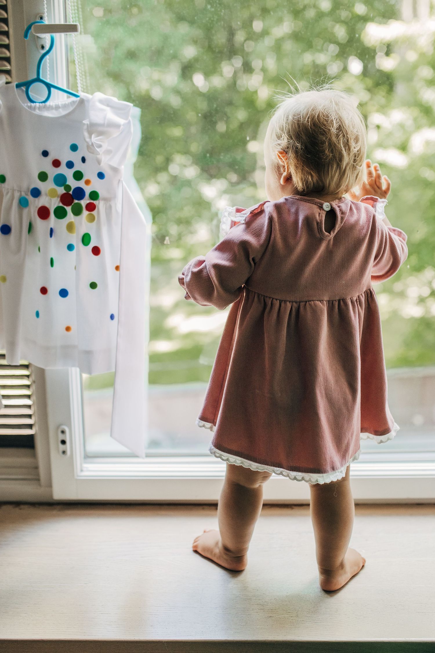 Toddler girl in pink dress standing at window beside colorful polka dot dress.