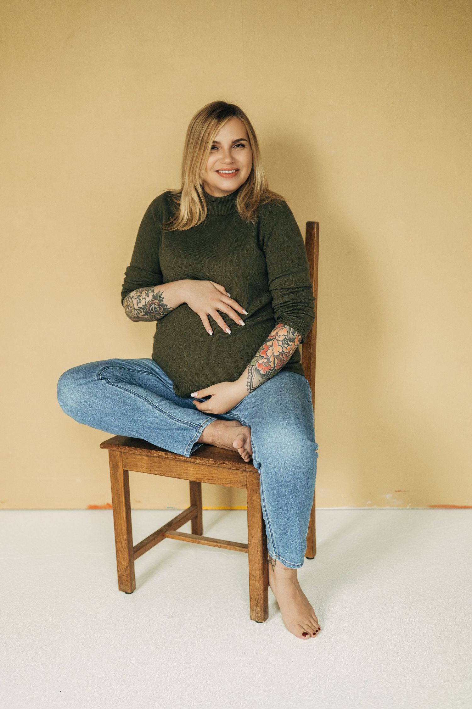 Pregnant woman in green sweater with tattoos sits cross-legged on wooden chair.
