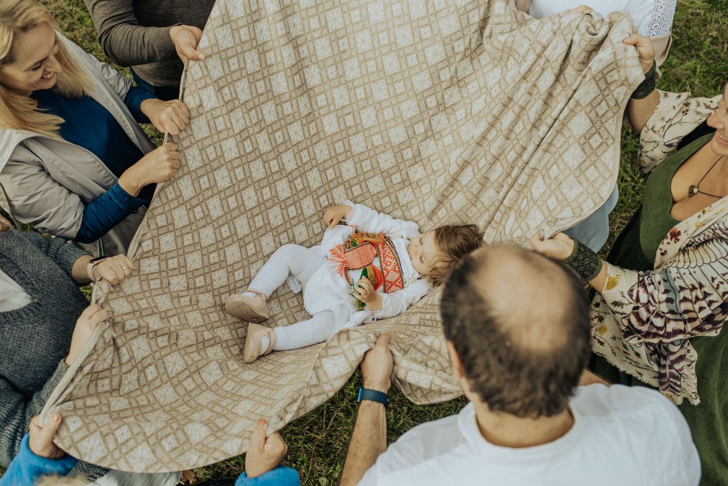 Group of people holding corners of a blanket with a baby lying in the center on green grass.