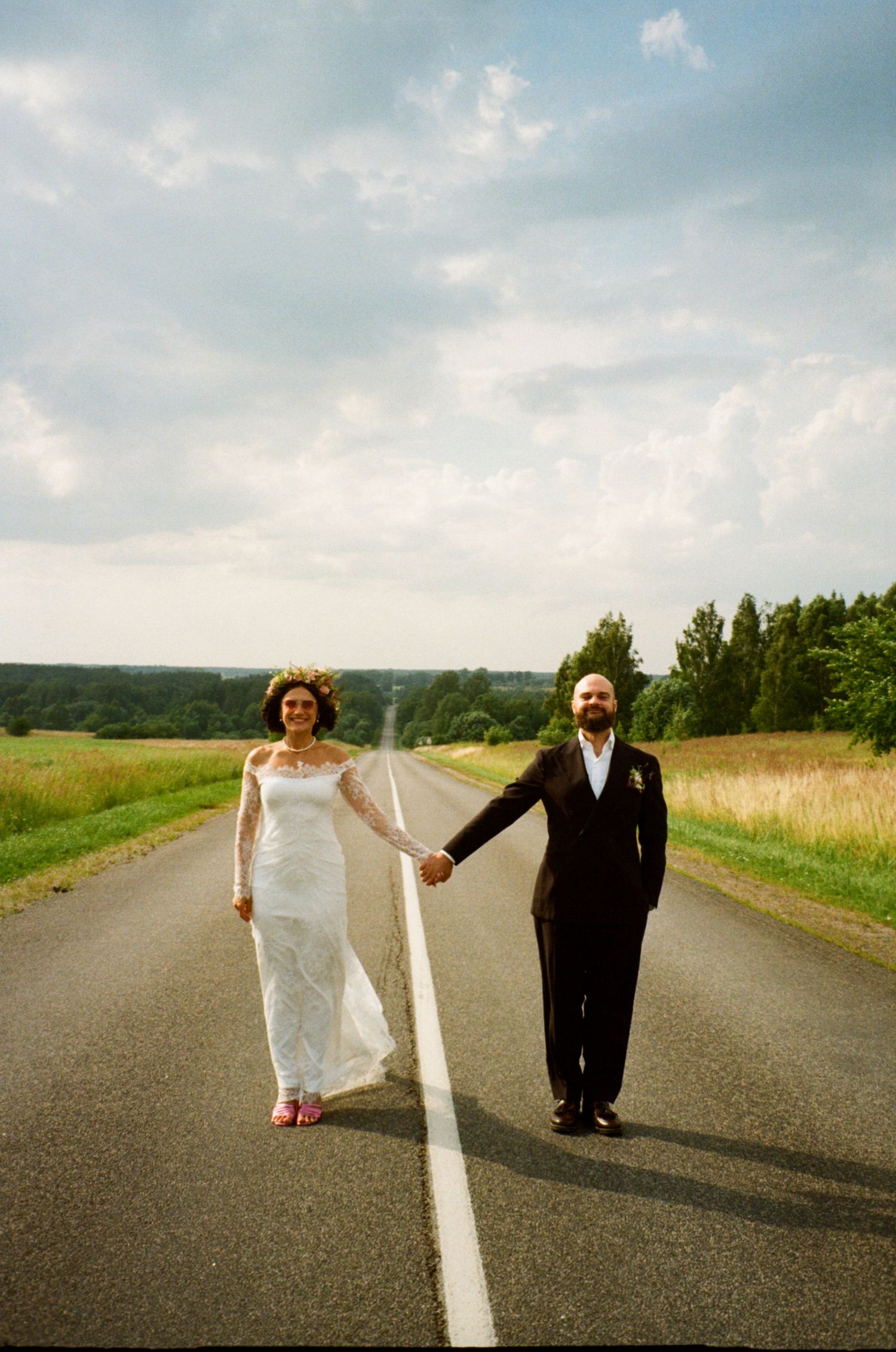 Bride and groom holding hands on an empty road surrounded by green fields and trees under a cloudy sky.