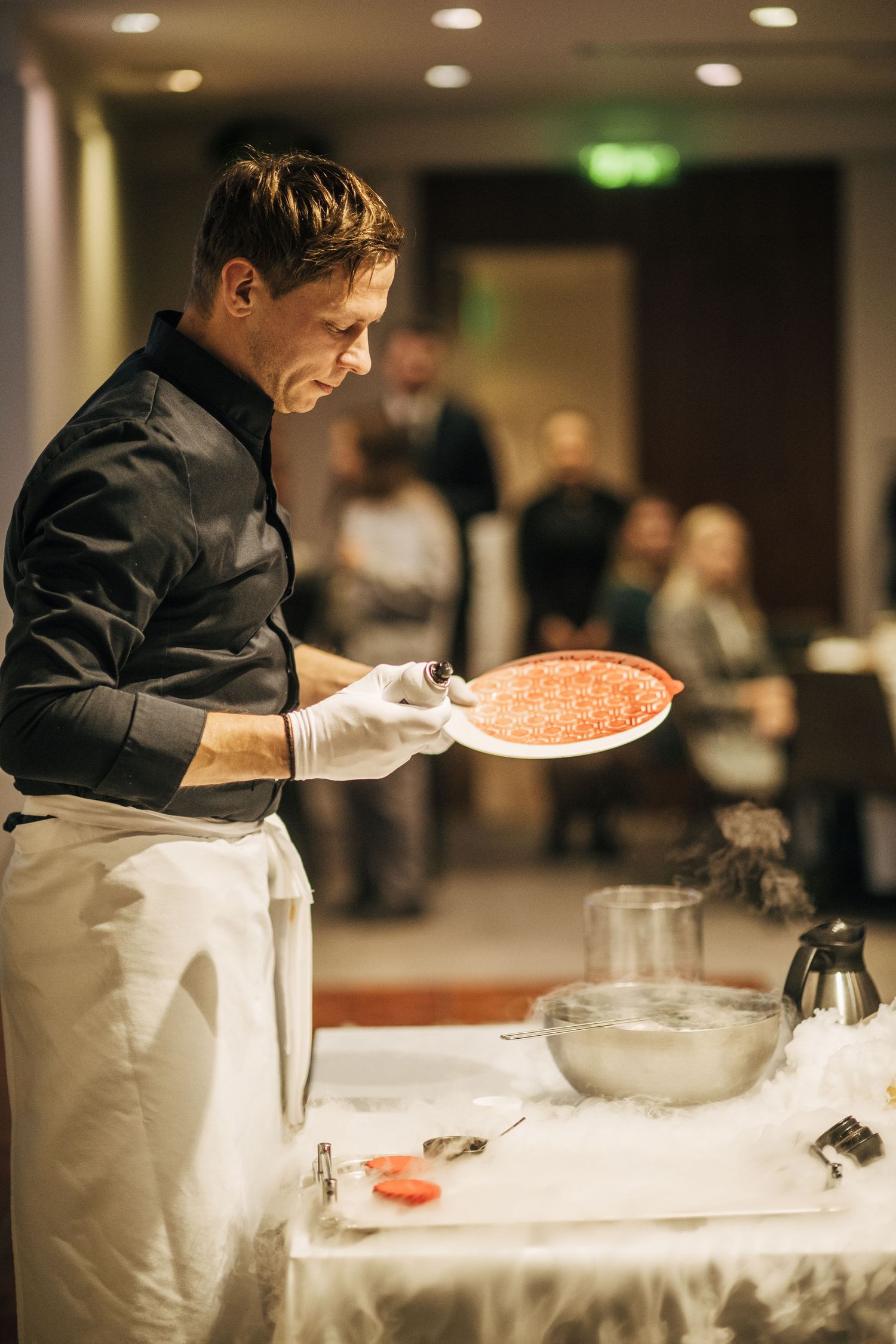 A chef in black uniform and white apron prepares food at a counter in a restaurant kitchen.