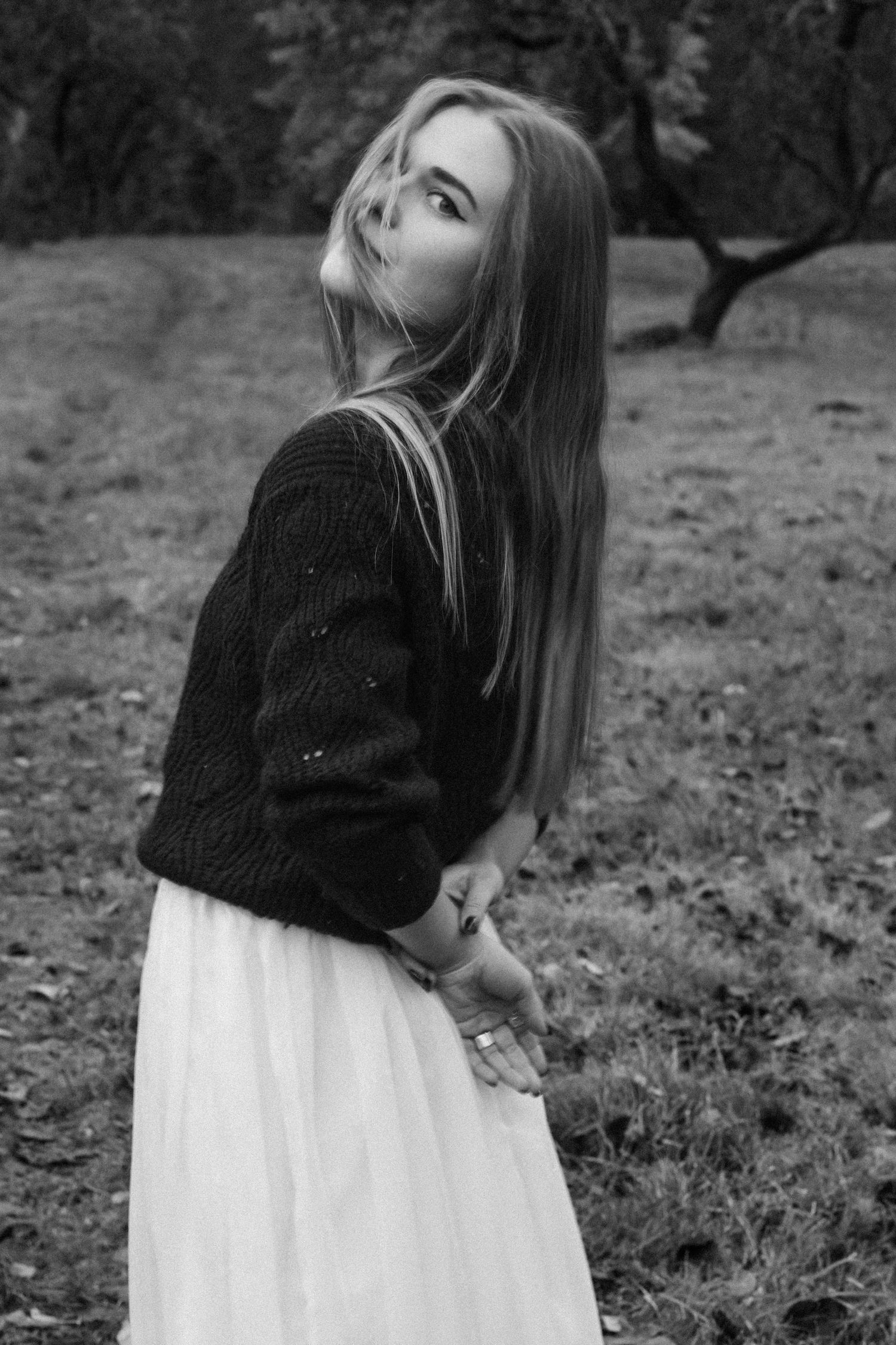 Black and white portrait of a person in a knit sweater and white skirt standing in a field looking up at the sky.