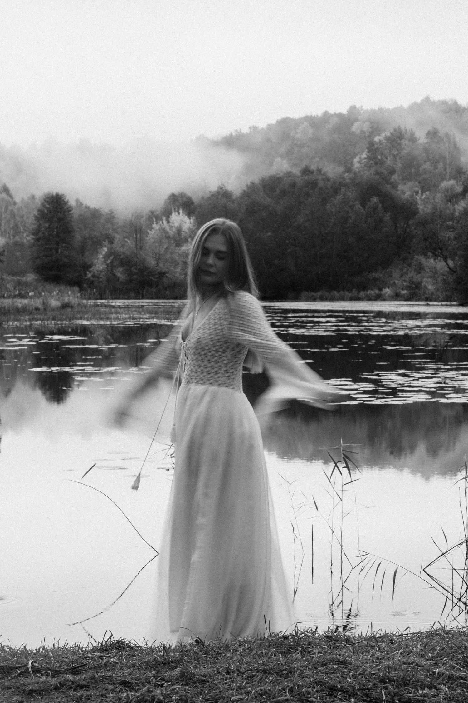 Ethereal black and white photo of a figure in a flowing white dress standing by a misty lake at dusk.