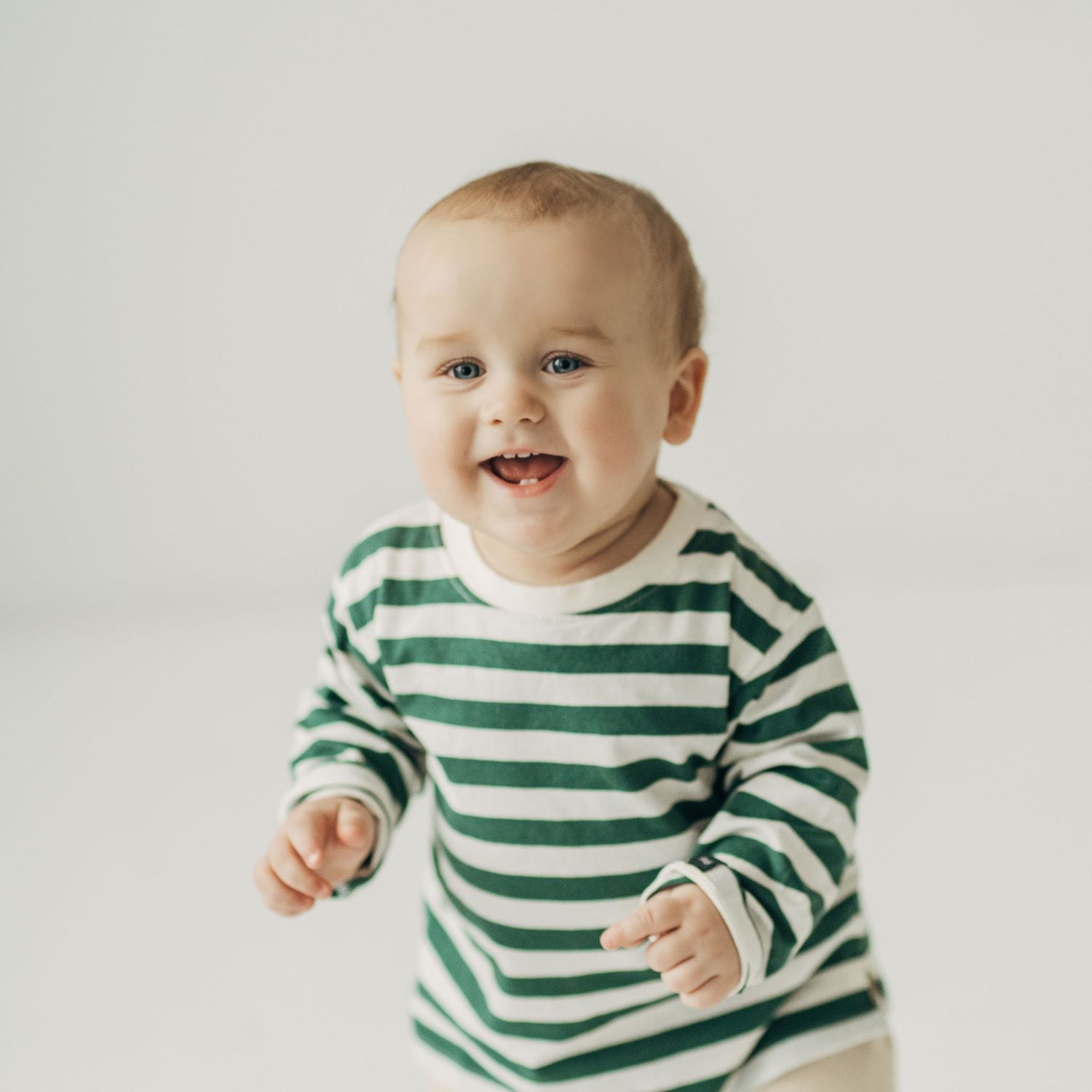 Baby in green striped shirt laughing and playing in a minimalist white setting.