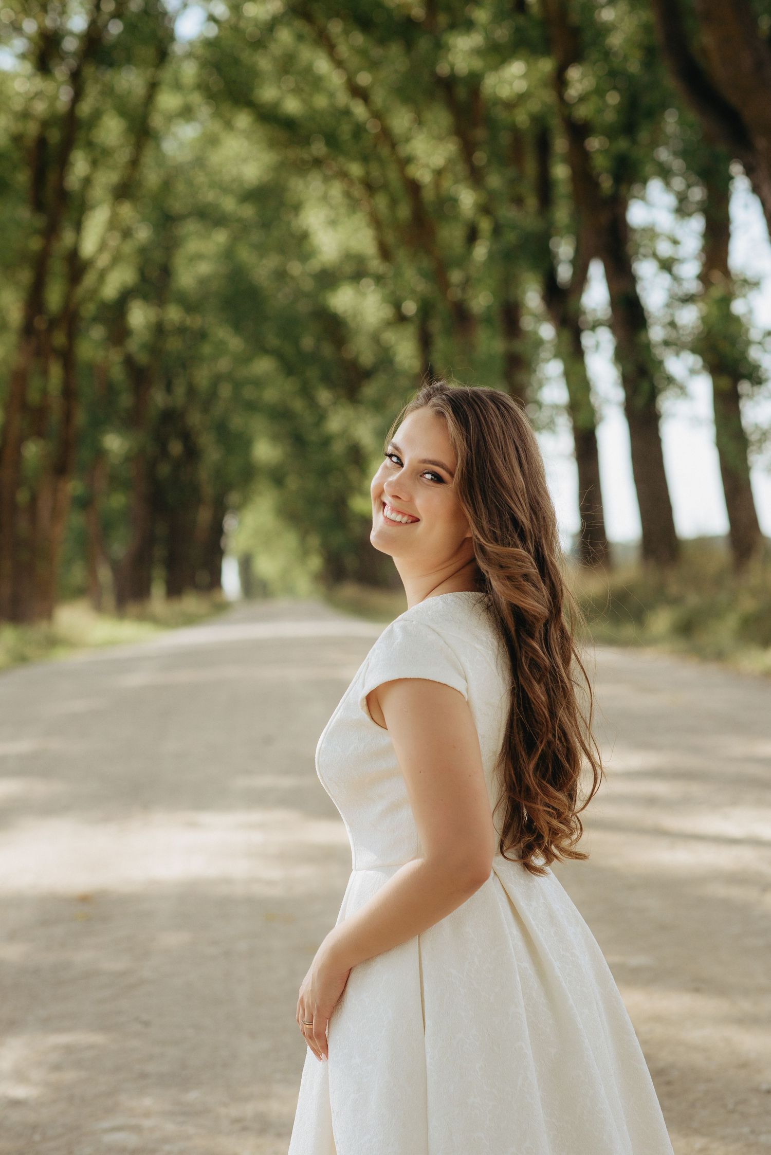 Bride in elegant white dress poses joyfully on a tree-lined path during golden hour.