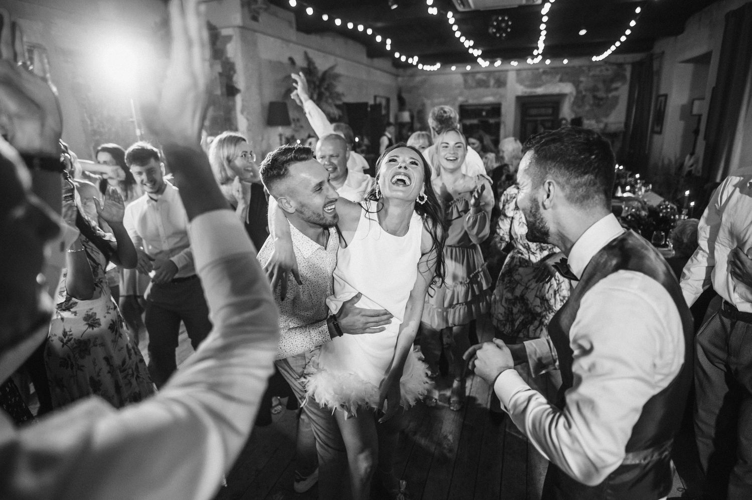 Energetic black and white photo of wedding reception dancing with string lights overhead.