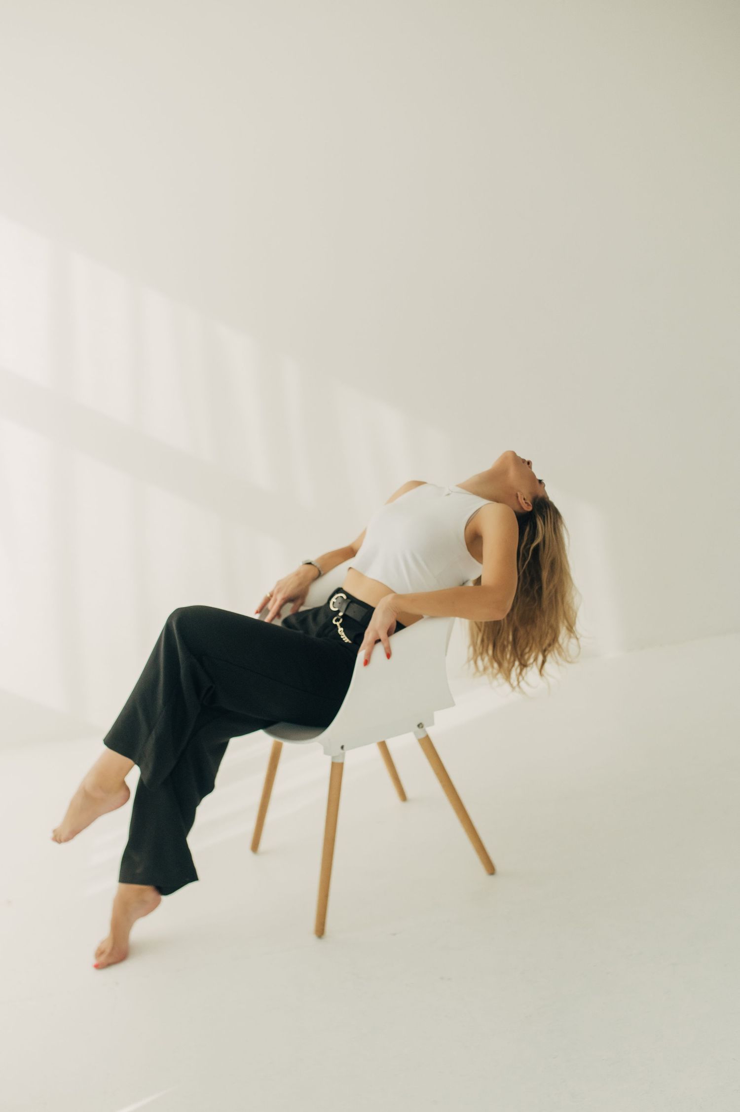 Person in white top and dark pants reclines on modern chair in minimalist studio setting.