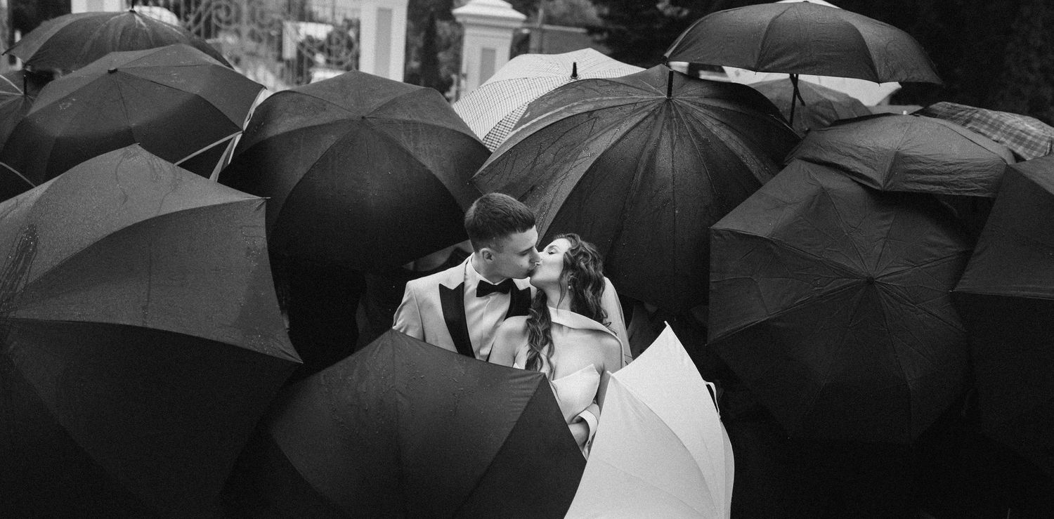 Black and white wedding photo of couple kissing surrounded by guests holding black umbrellas in the rain.