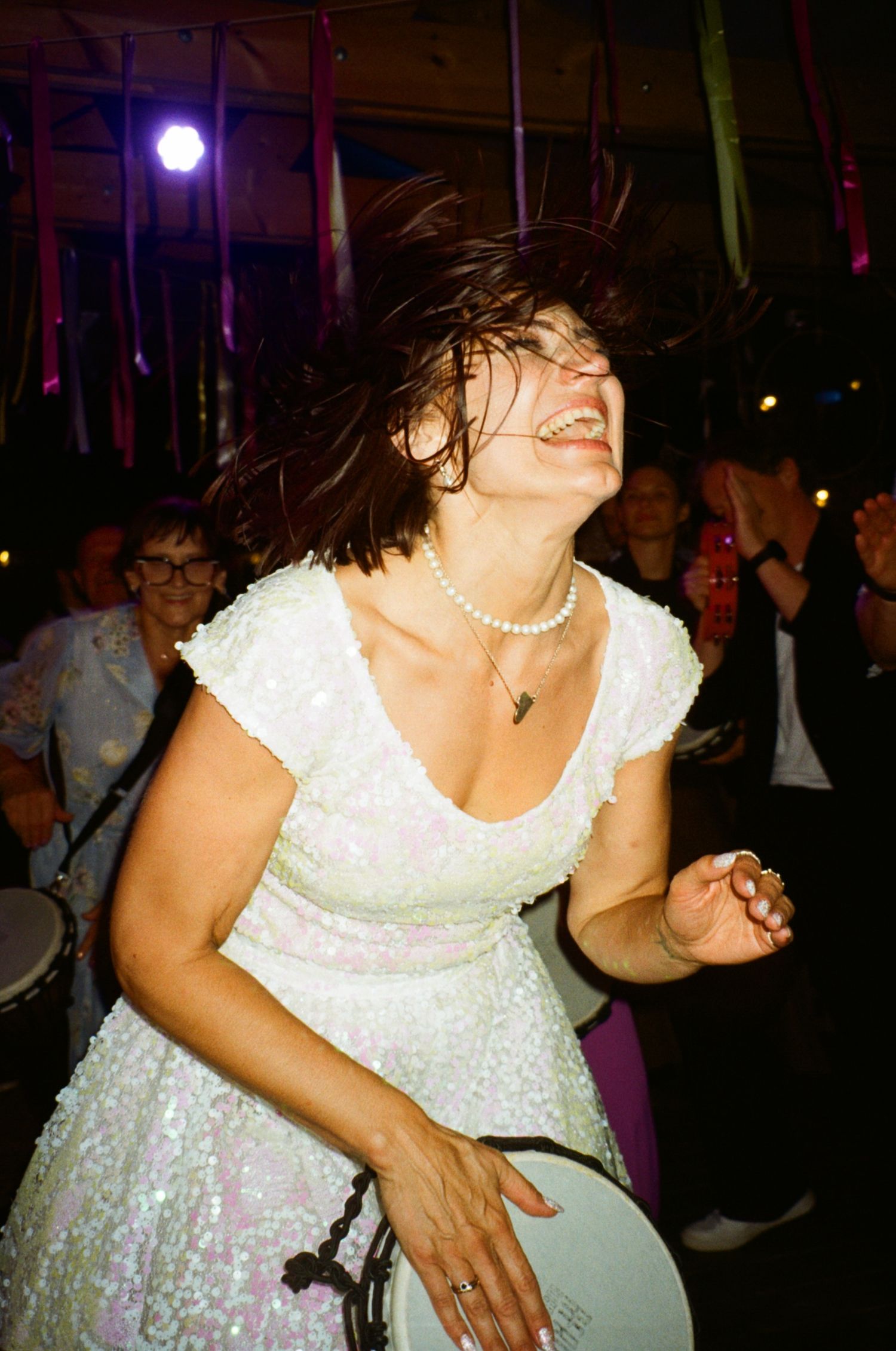 A joyful woman in a white sequin dress dances energetically, her hair flying, wearing a pearl necklace at a party.