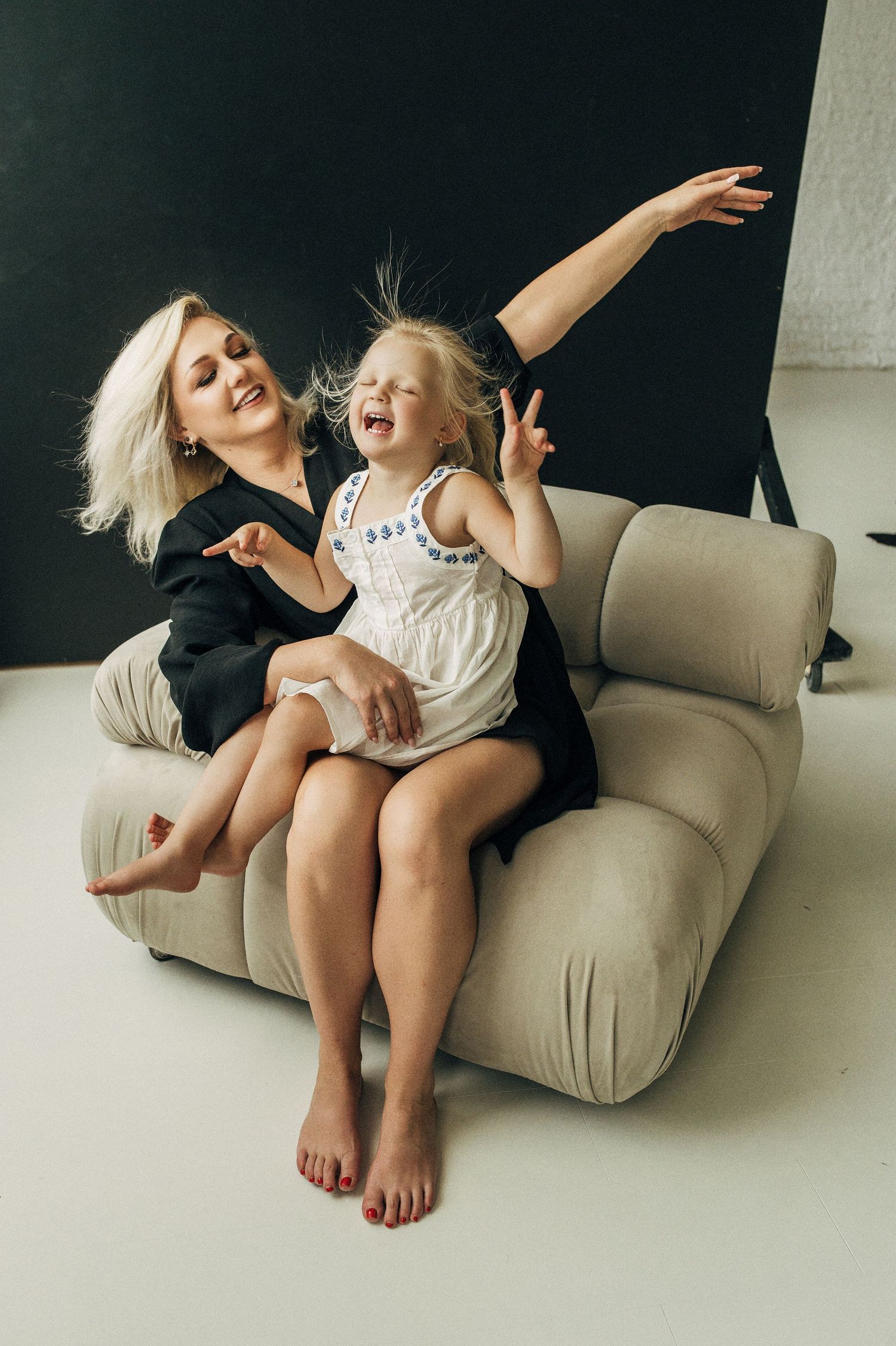 Two people in casual attire share playful moments while lounging on a beige couch in a minimalist setting.