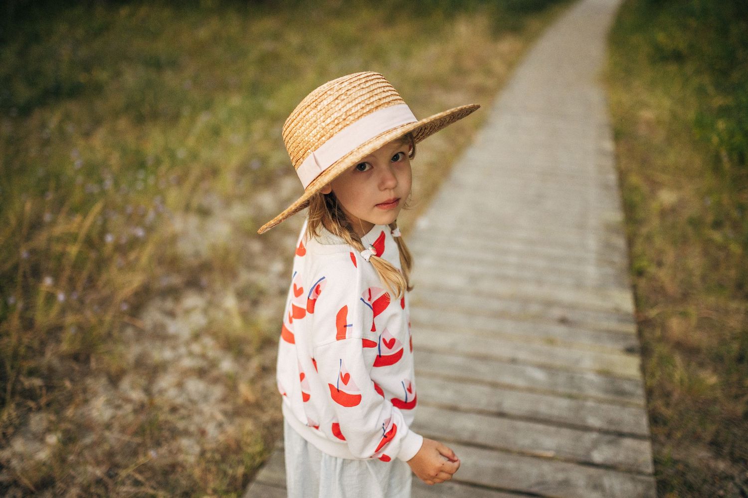 Young girl in straw hat and floral top stands on wooden boardwalk path surrounded by tall grass.