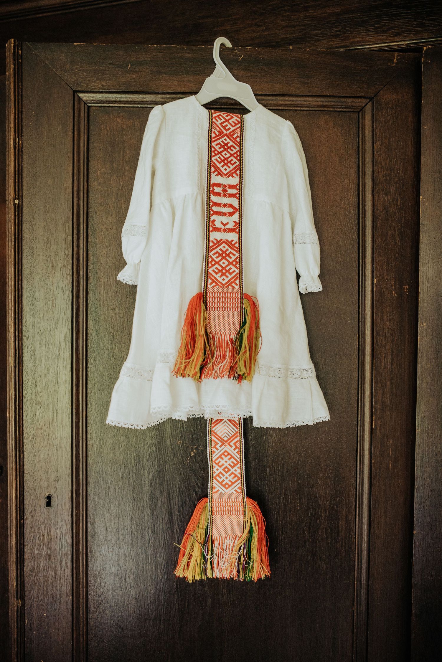White embroidered christening gown with colorful traditional woven sash on wooden door.