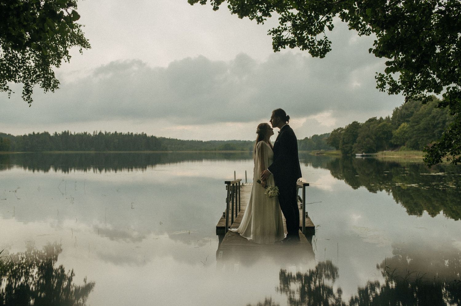 A wedding couple stands by a serene lake with fog rolling across the water and trees in the background.
