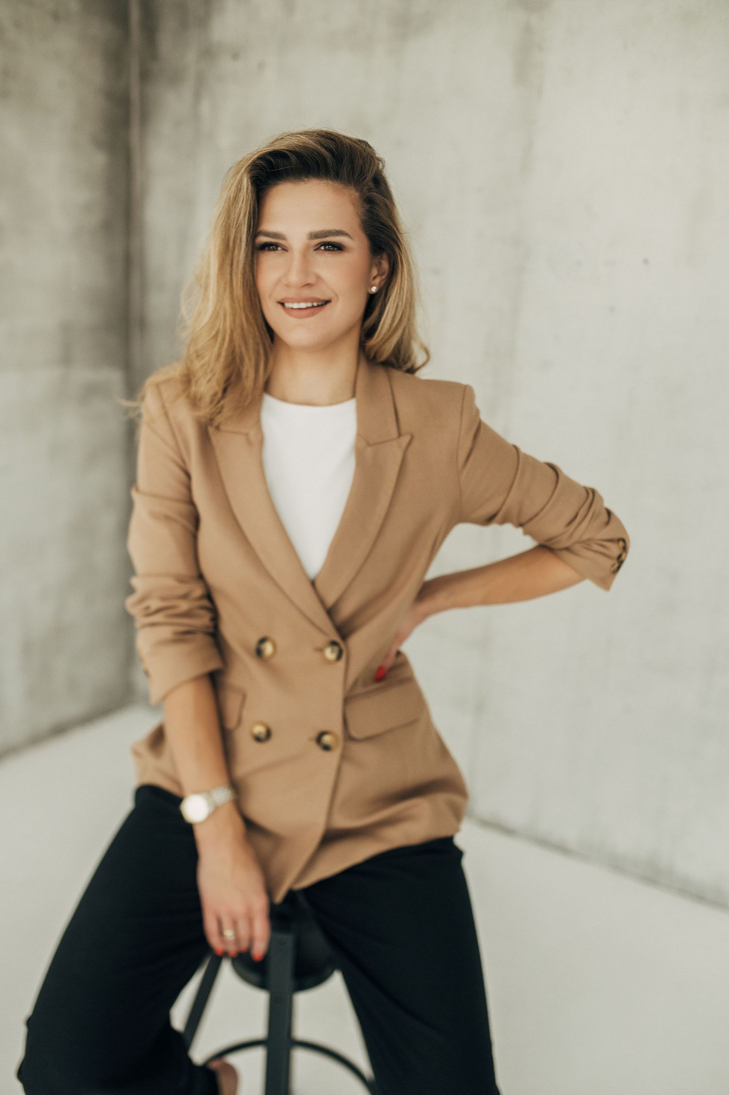 Person in tan blazer and white shirt posing on chair in studio setting.