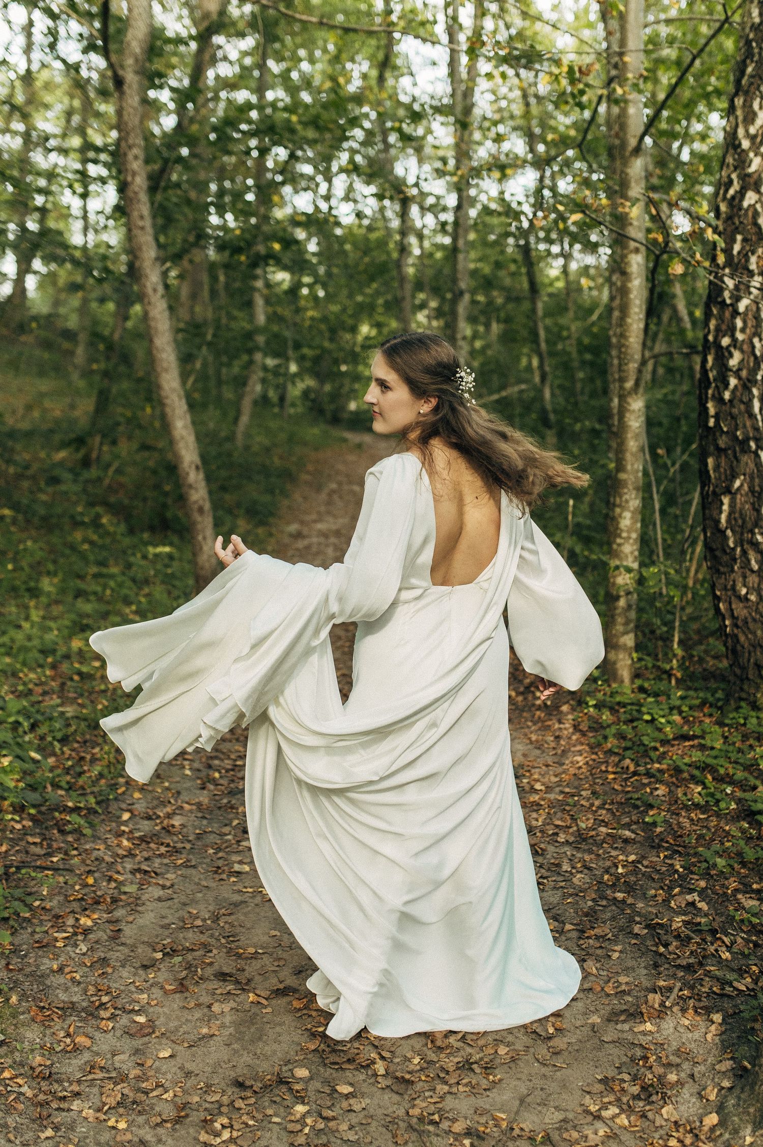 A flowing white backless dress with billowing sleeves twirls gracefully on a forest path among tall trees.