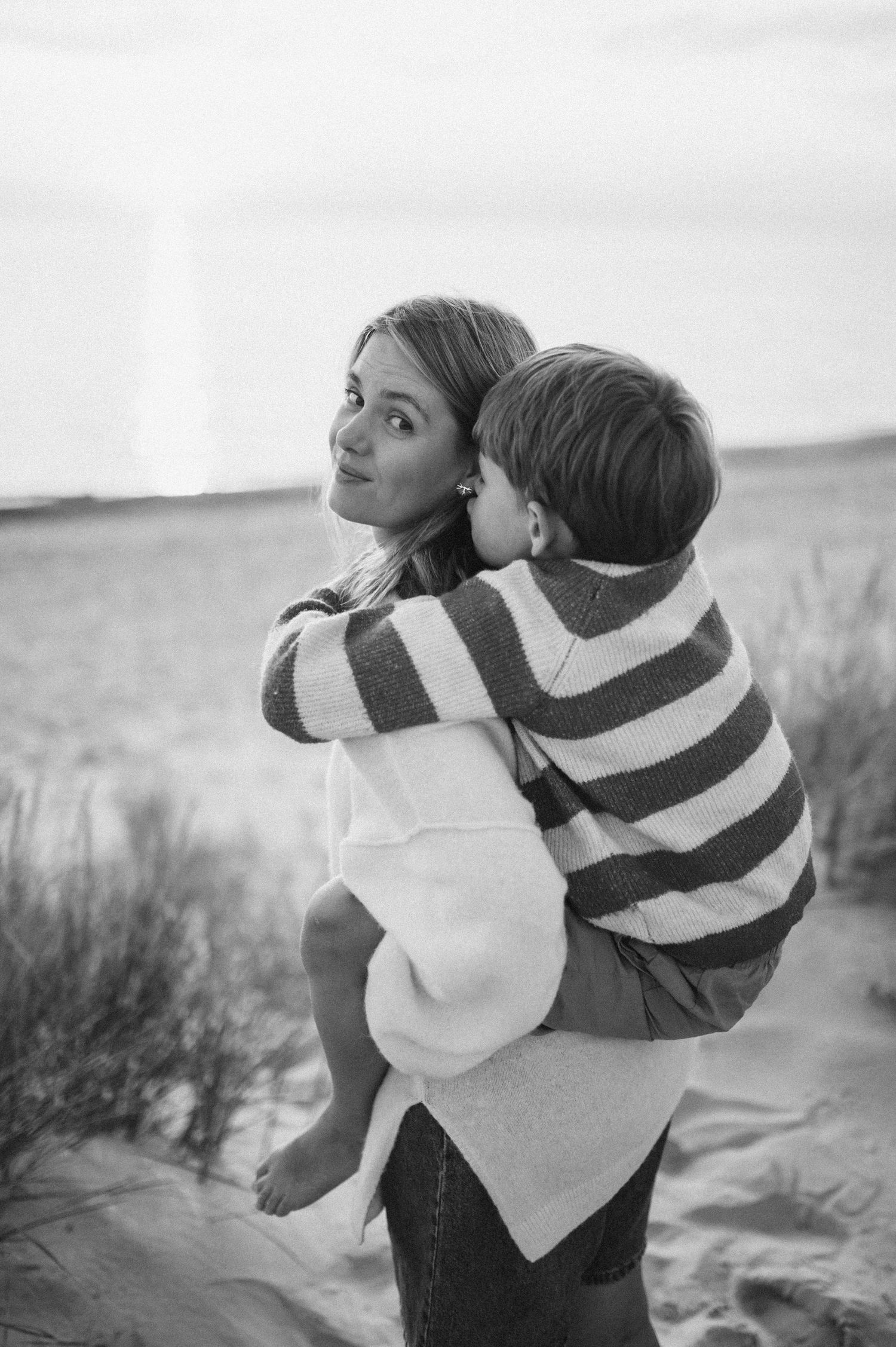 Black and white photo of a woman holding a child in a striped sweater on a sandy beach.