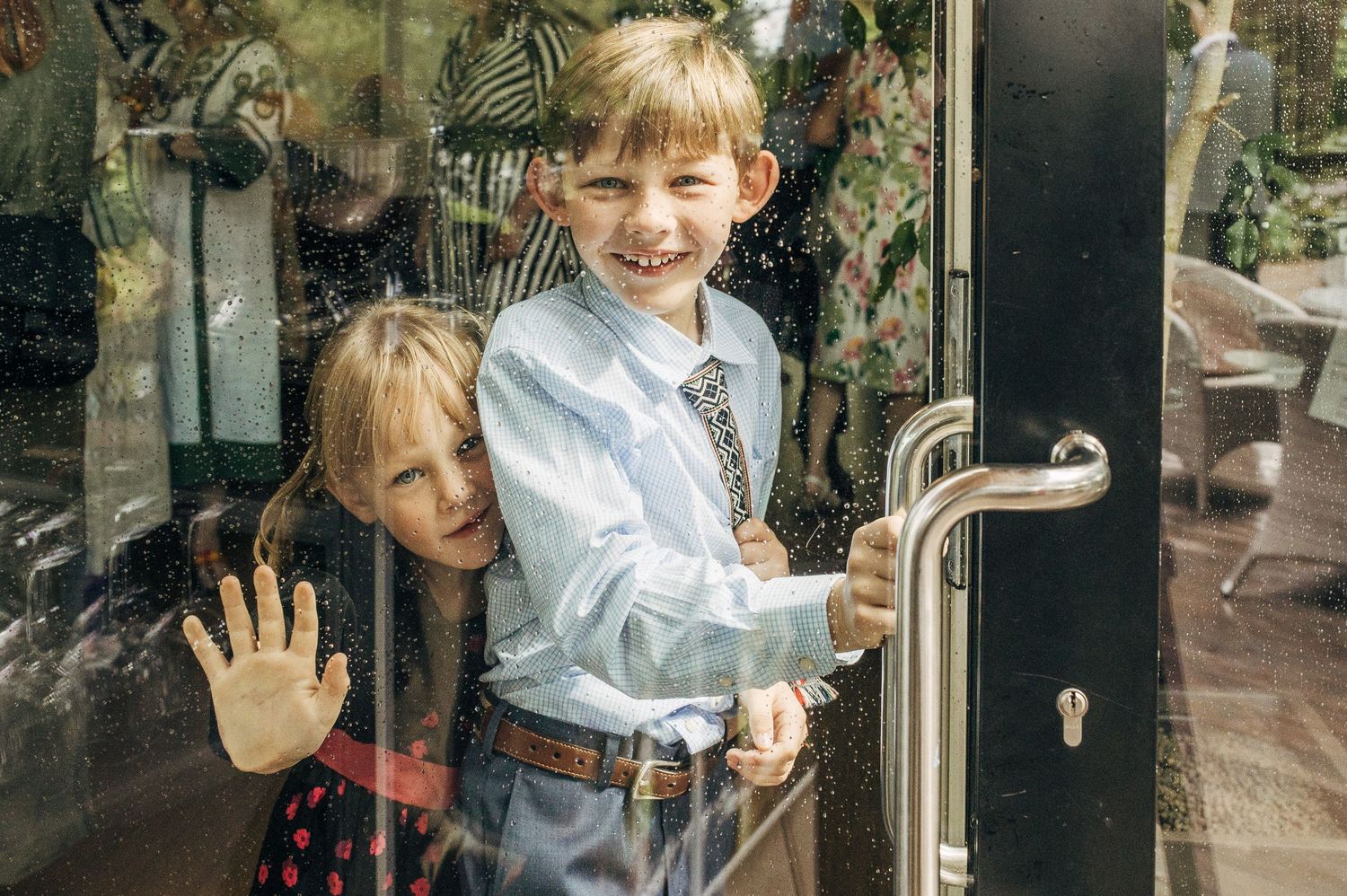 Two happy children peeking and waving through a glass door at a formal event.