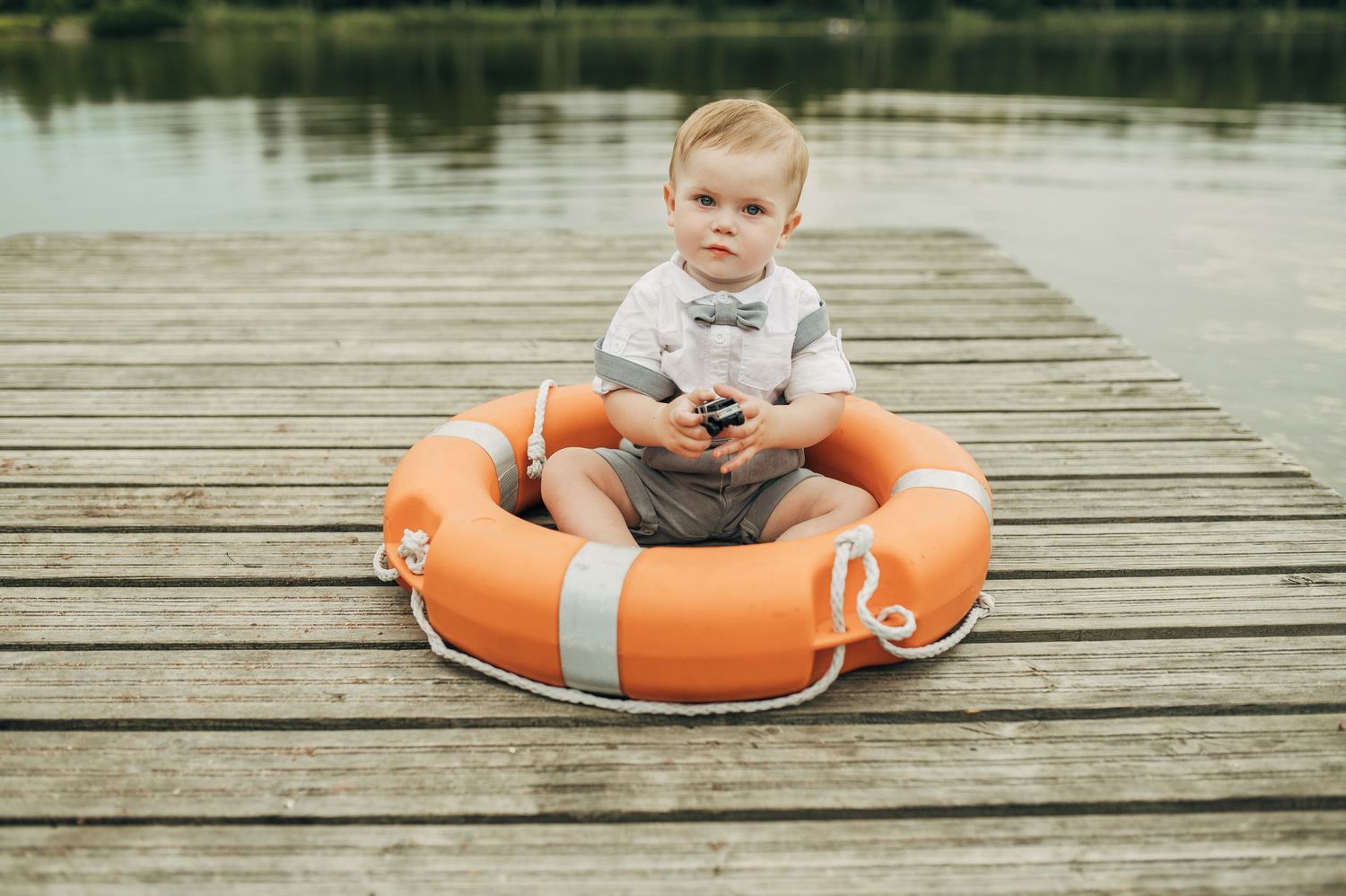 Cute baby boy sitting inside an orange life preserver ring on a wooden dock by a lake.