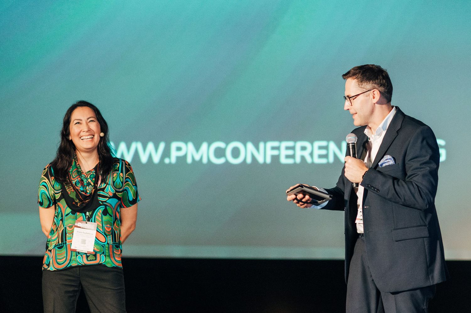 Speakers presenting at a professional conference with teal backdrop and PM Conference branding.
