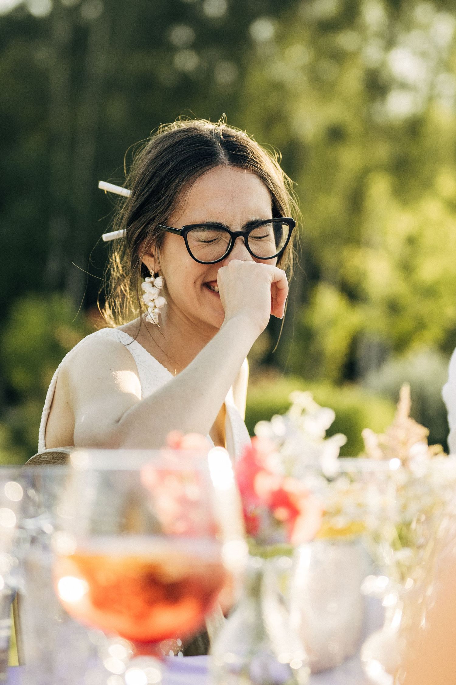 Young woman with glasses laughing at an outdoor garden party, seated at a decorated table with floral arrangements.