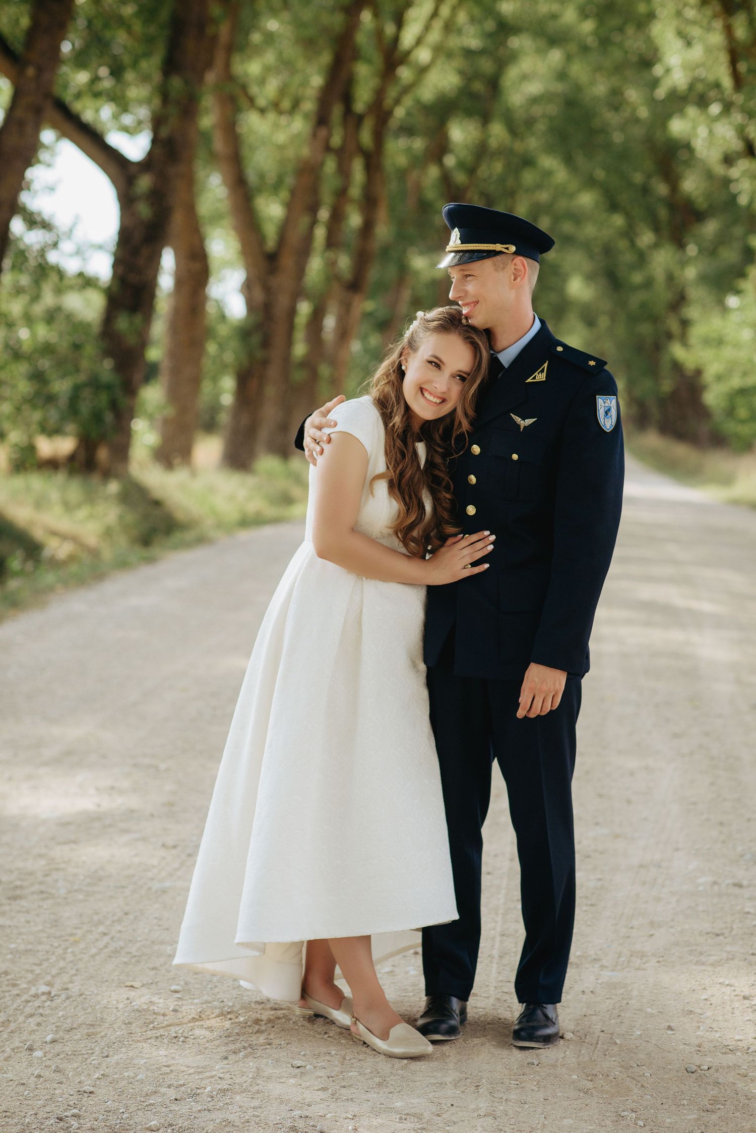 Two young children in formal wear share a sweet moment on a tree-lined path.