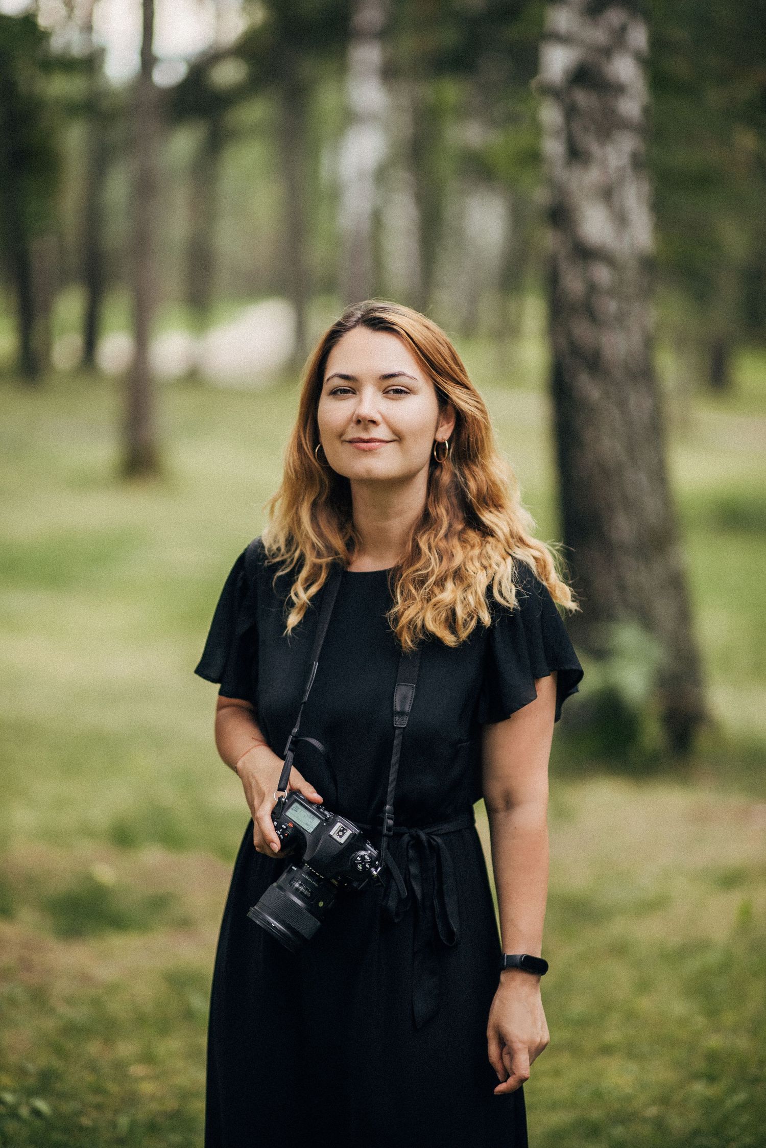 Wedding photographer in black dress with camera equipment in forest setting.