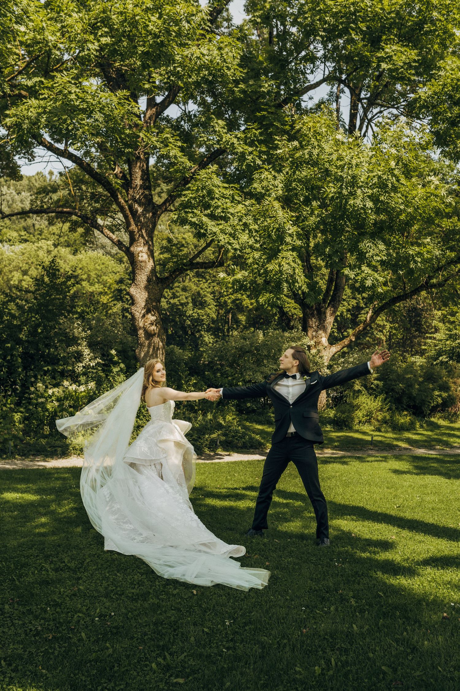 Bride in white gown and groom in black tuxedo dancing joyfully on green lawn surrounded by lush trees.