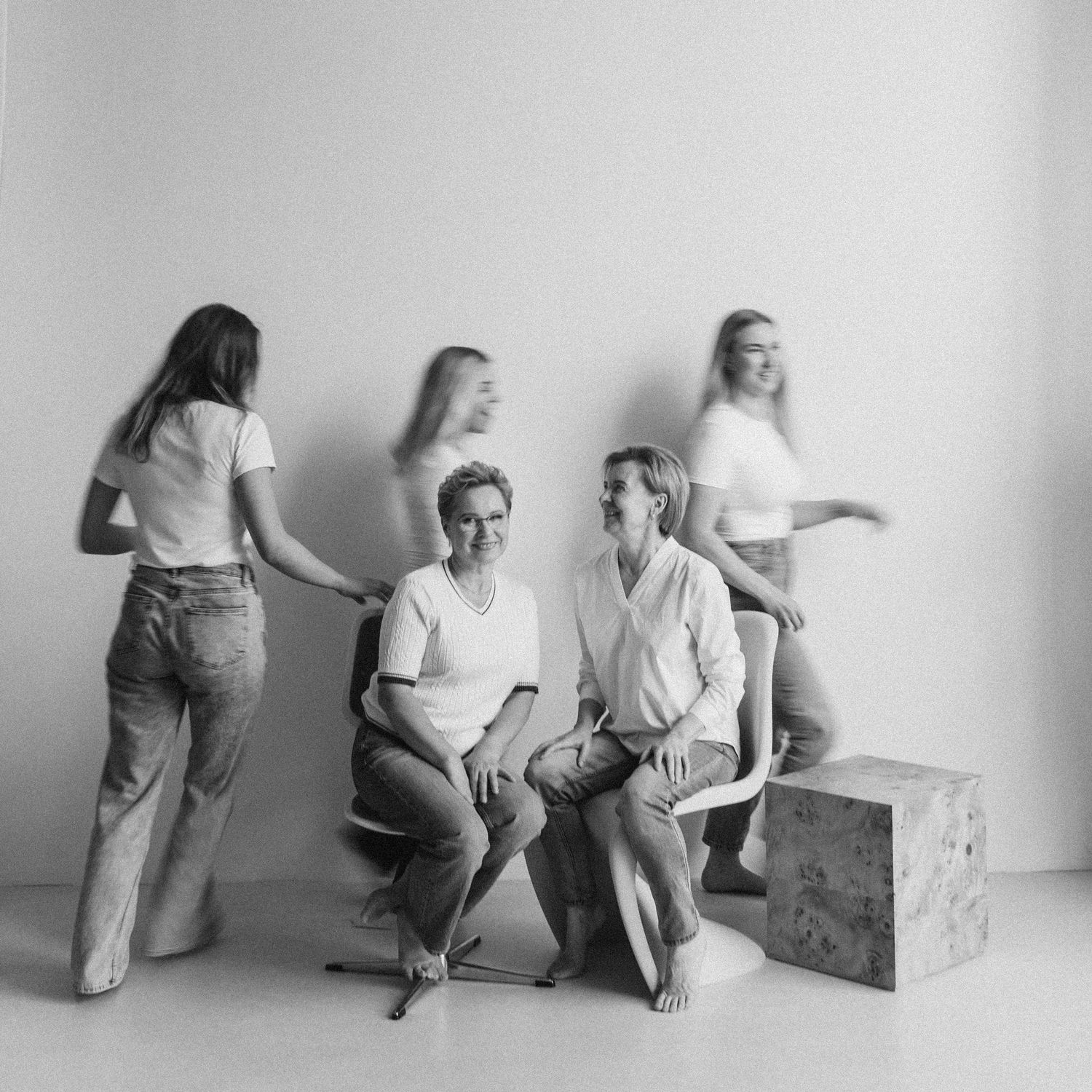 Black and white photo of two people sitting on chairs while two others move around them in a bright studio.