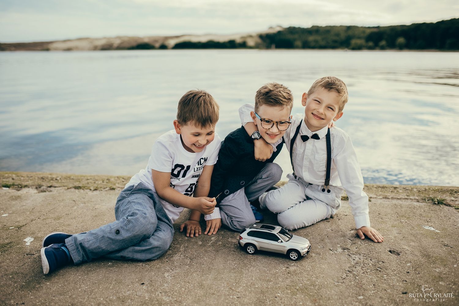 Three young boys playing with a toy car on a sandy beach near calm water, dressed in smart casual outfits.