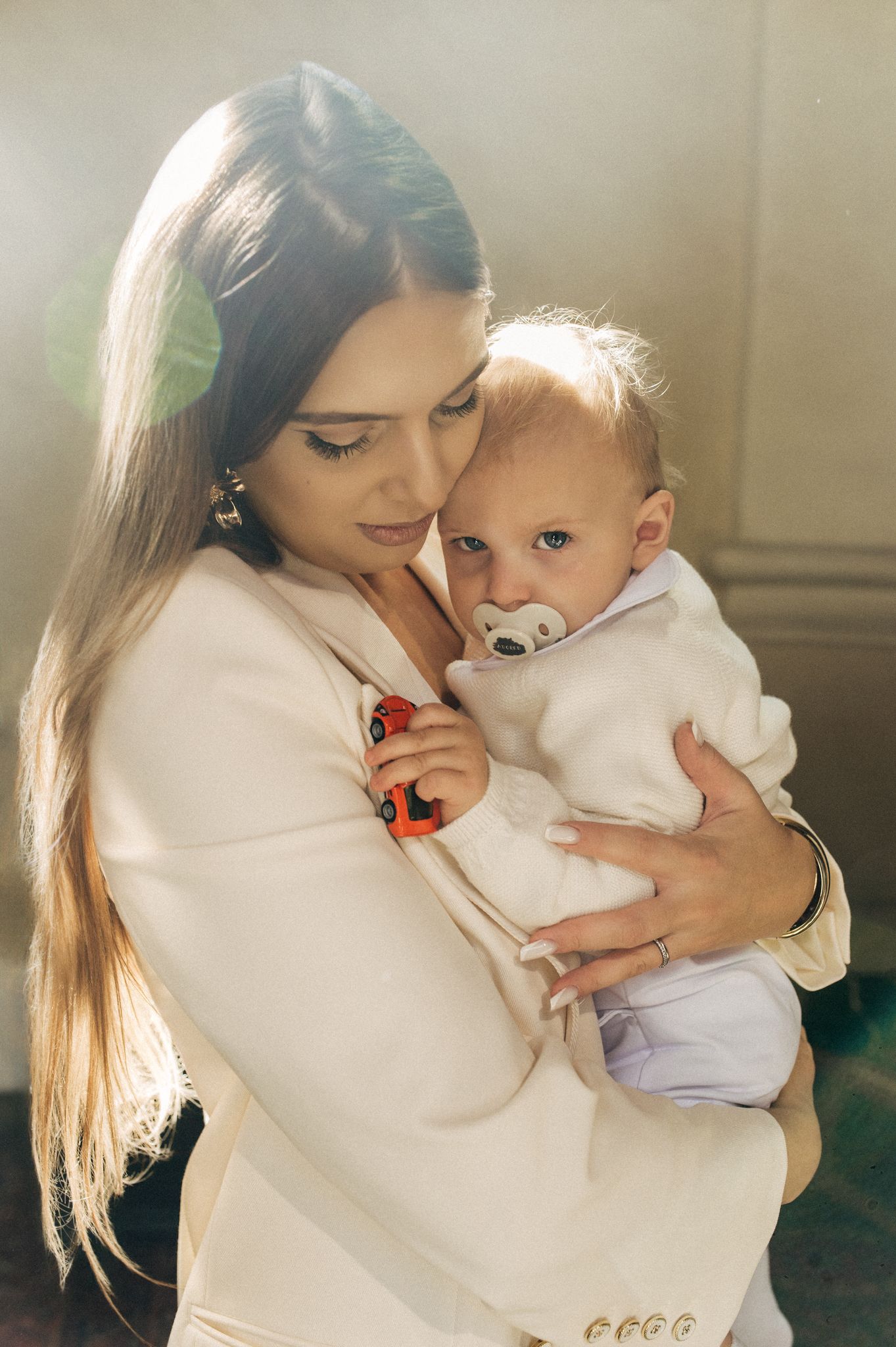 A young mother in a white blazer tenderly holds her baby with a pacifier indoors.