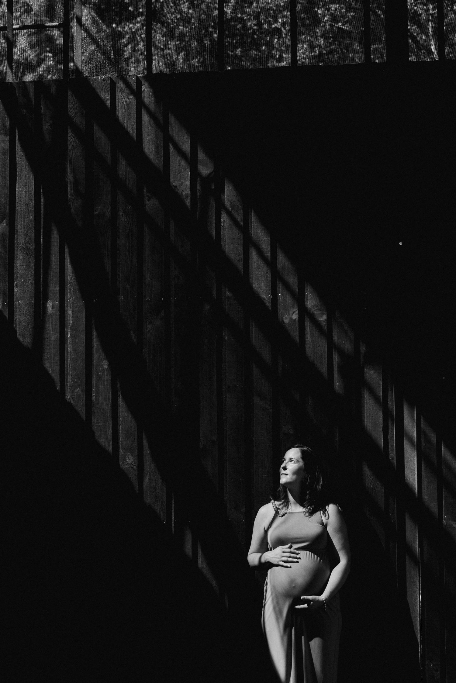 Dramatic black and white photo of pregnant woman standing in shadows with sunlight streaming through.