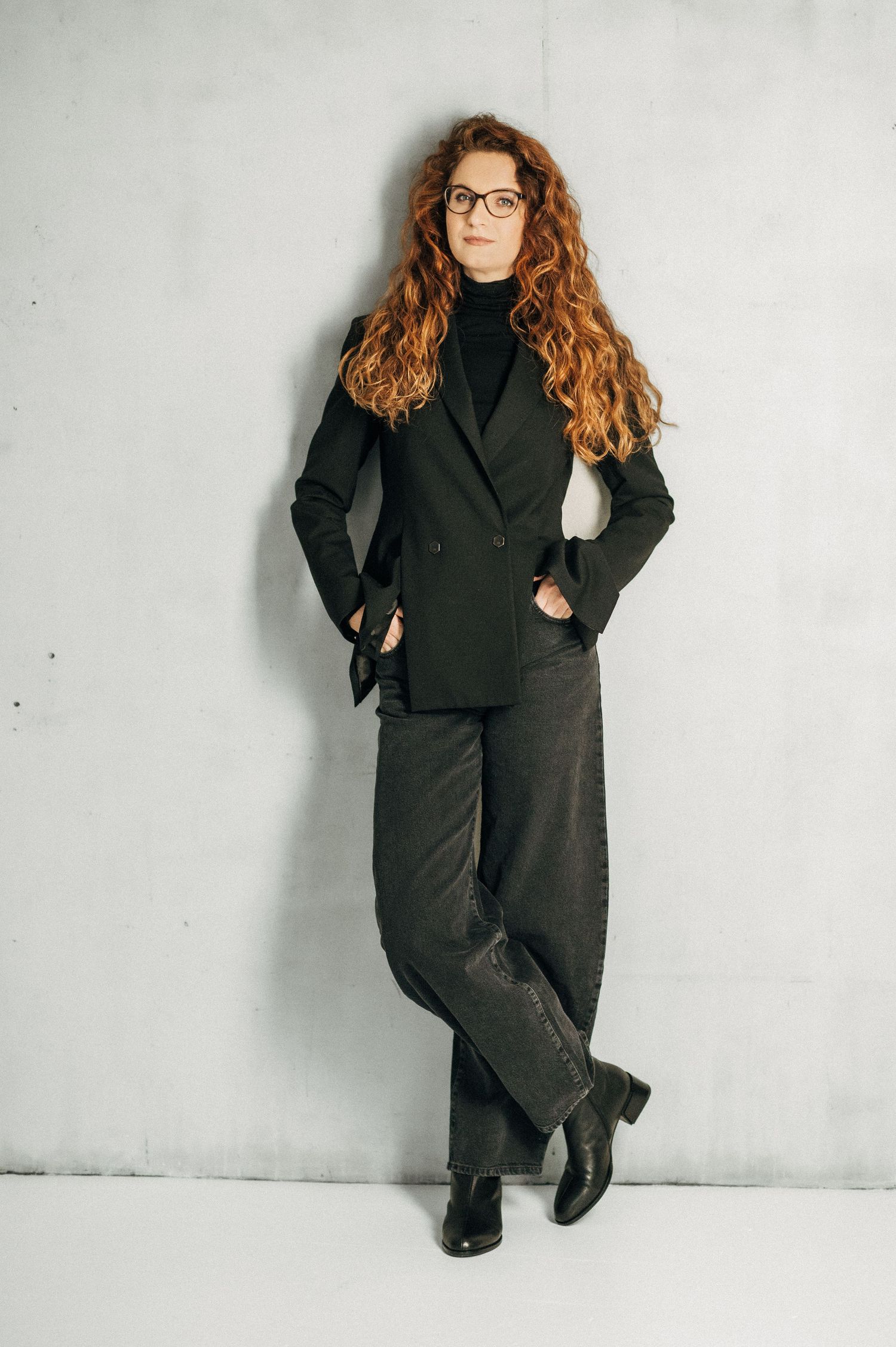 A person in a black business suit and glasses poses against a white wall in a series of professional fashion shots.