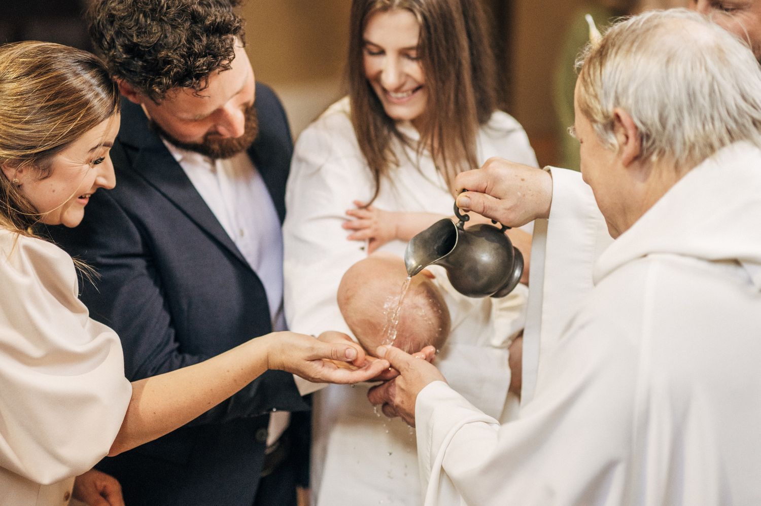 Priest performing a baby baptism ceremony with parents and godparents in a church.