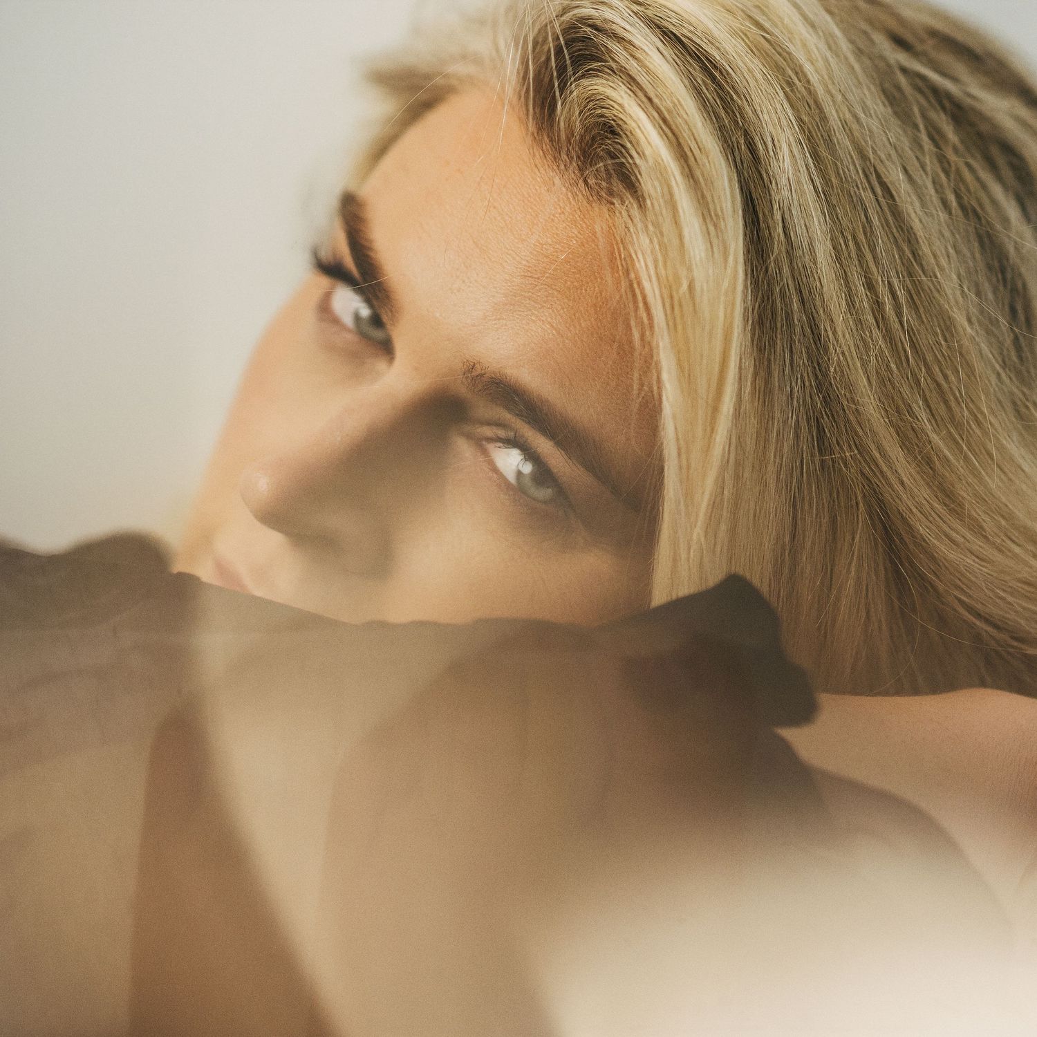 A blonde woman rests her chin on her arms, gazing upward with soft natural lighting creating elegant shadows.