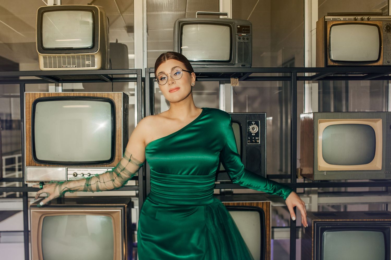 Stylish poses against vintage television wall wearing emerald green evening dress.