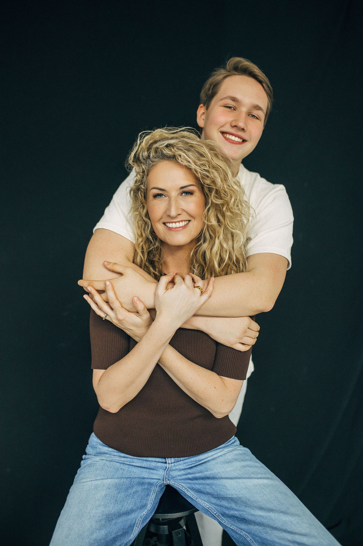 A young couple smiling, man hugging woman from behind, both in casual attire against a dark background.