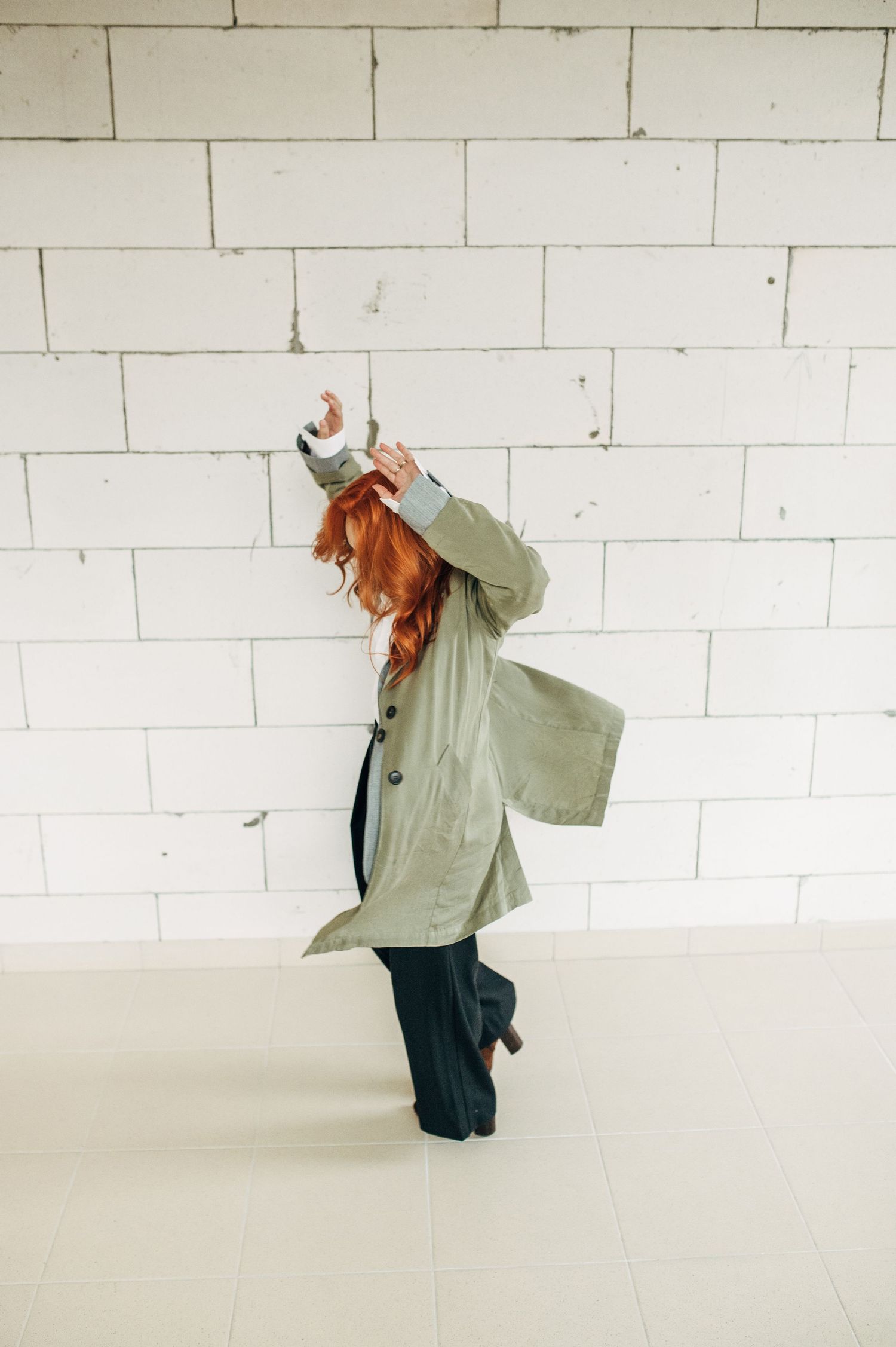 Figure in light jacket moves expressively against white brick wall in studio.