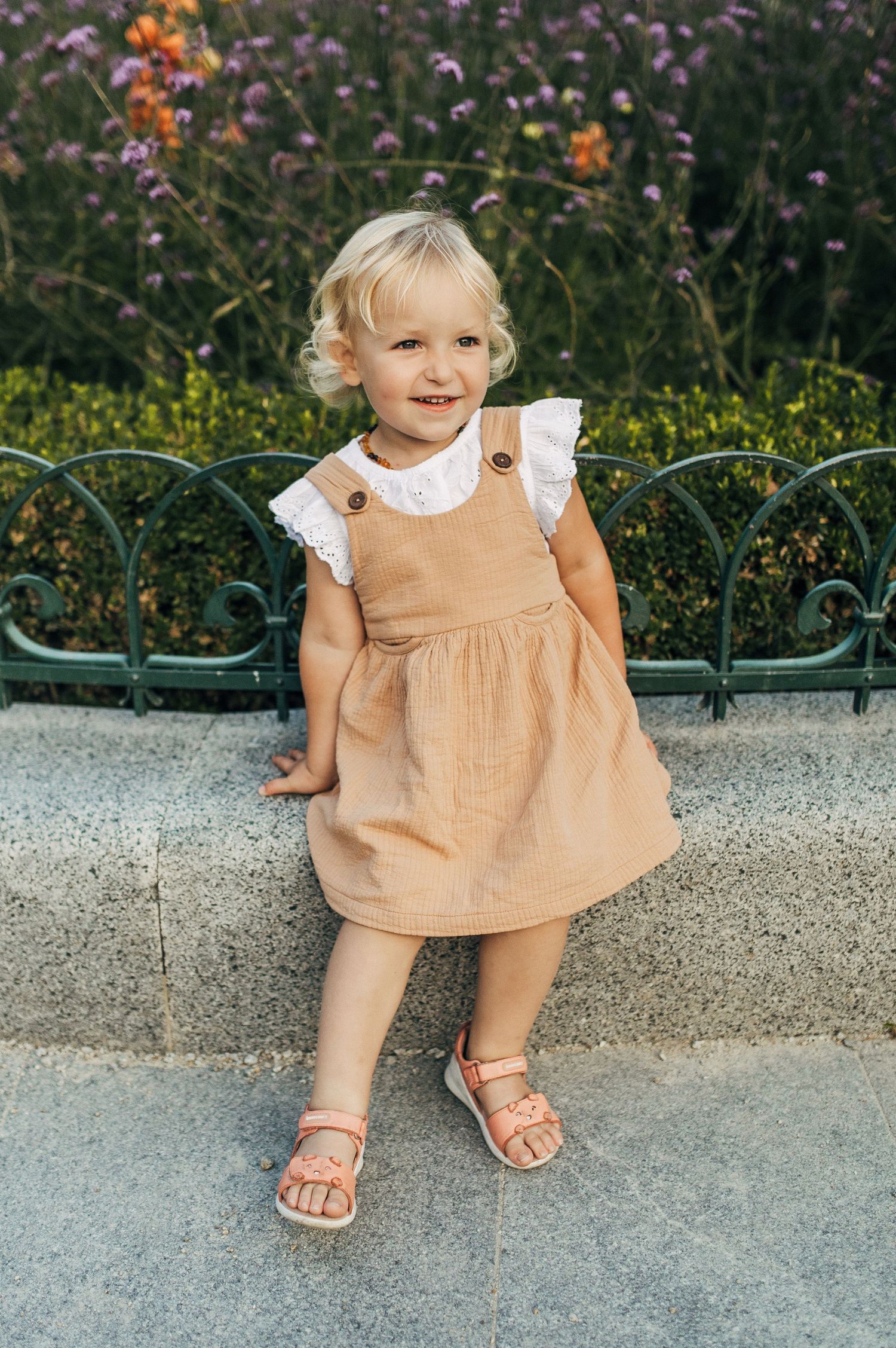 Close-up of small feet in sandals and peachy dress against colorful garden flowers.