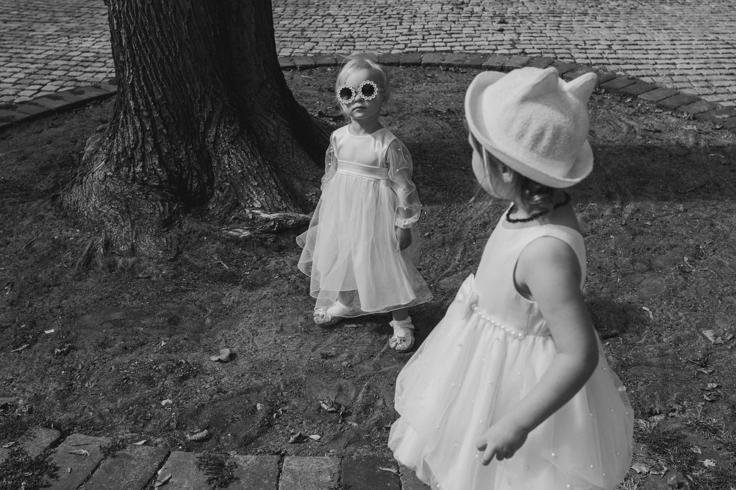Child in white dress plays joyfully outside near trees in black and white photography.